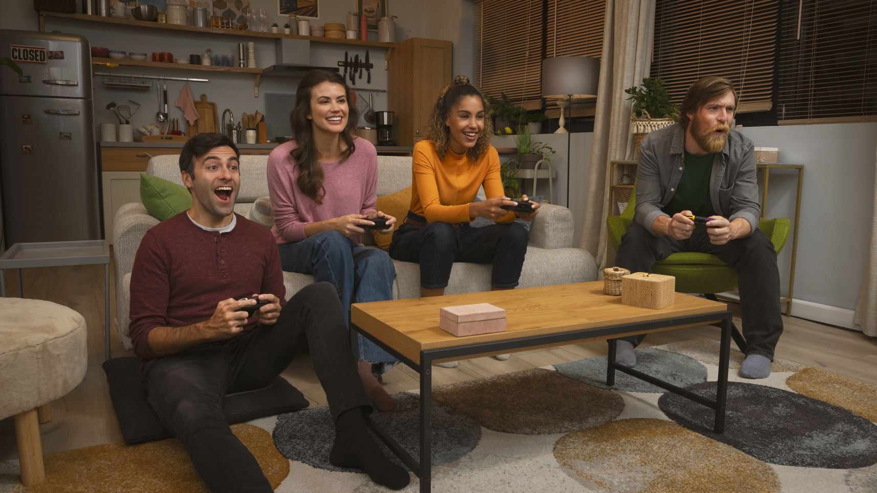 Four people playing video games, sitting on a couch and floor, smiling.
