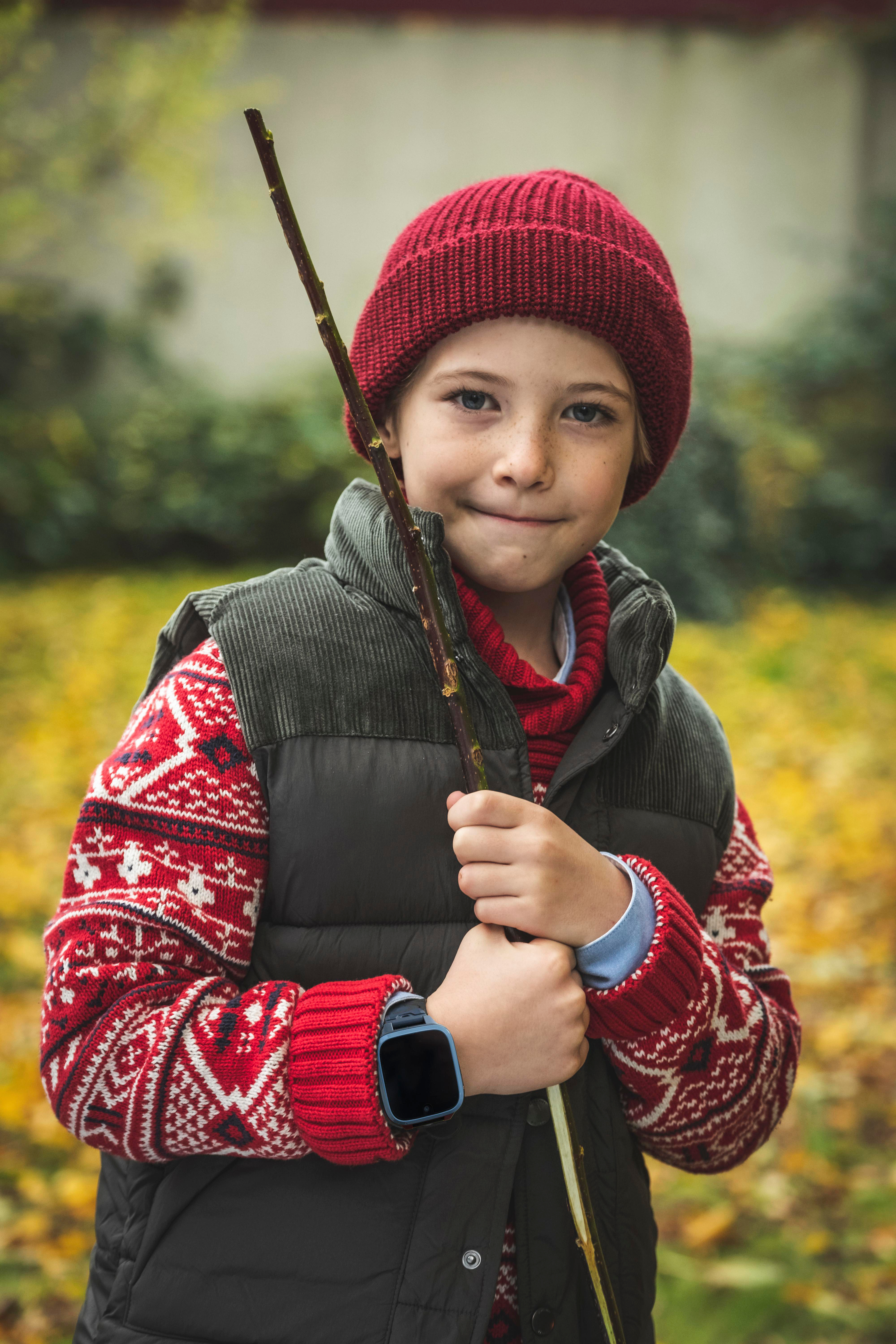 Ein Junge mit roter Mütze und Pullover, der einen Stock hält, lächelt in einem Park.