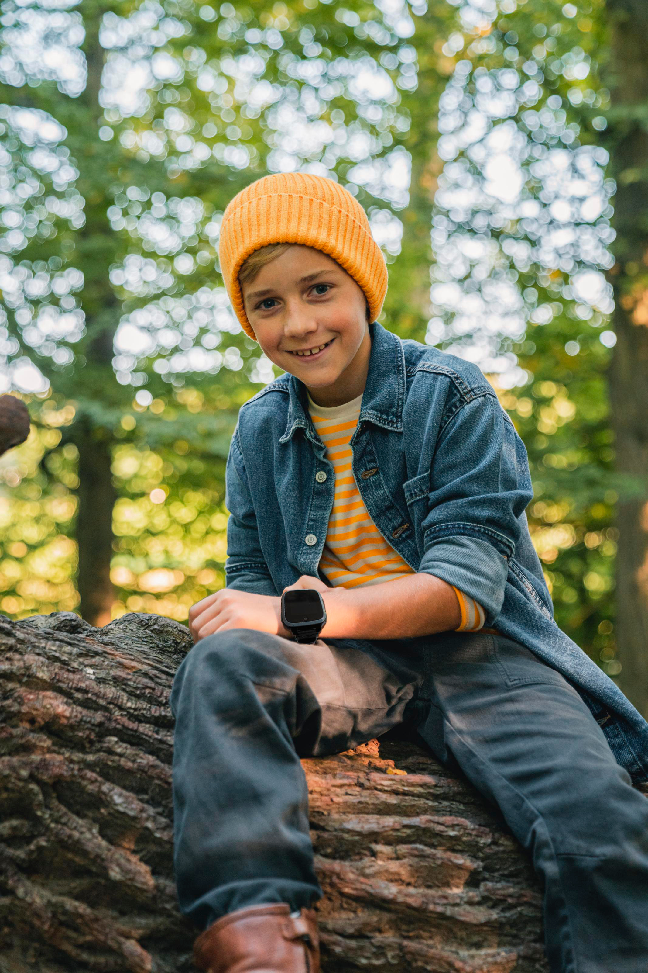 Junge mit orangefarbener Mütze und Jeansjacke sitzt auf einem Baumstamm im Wald. Lächelt, hält eine schwarze Uhr.