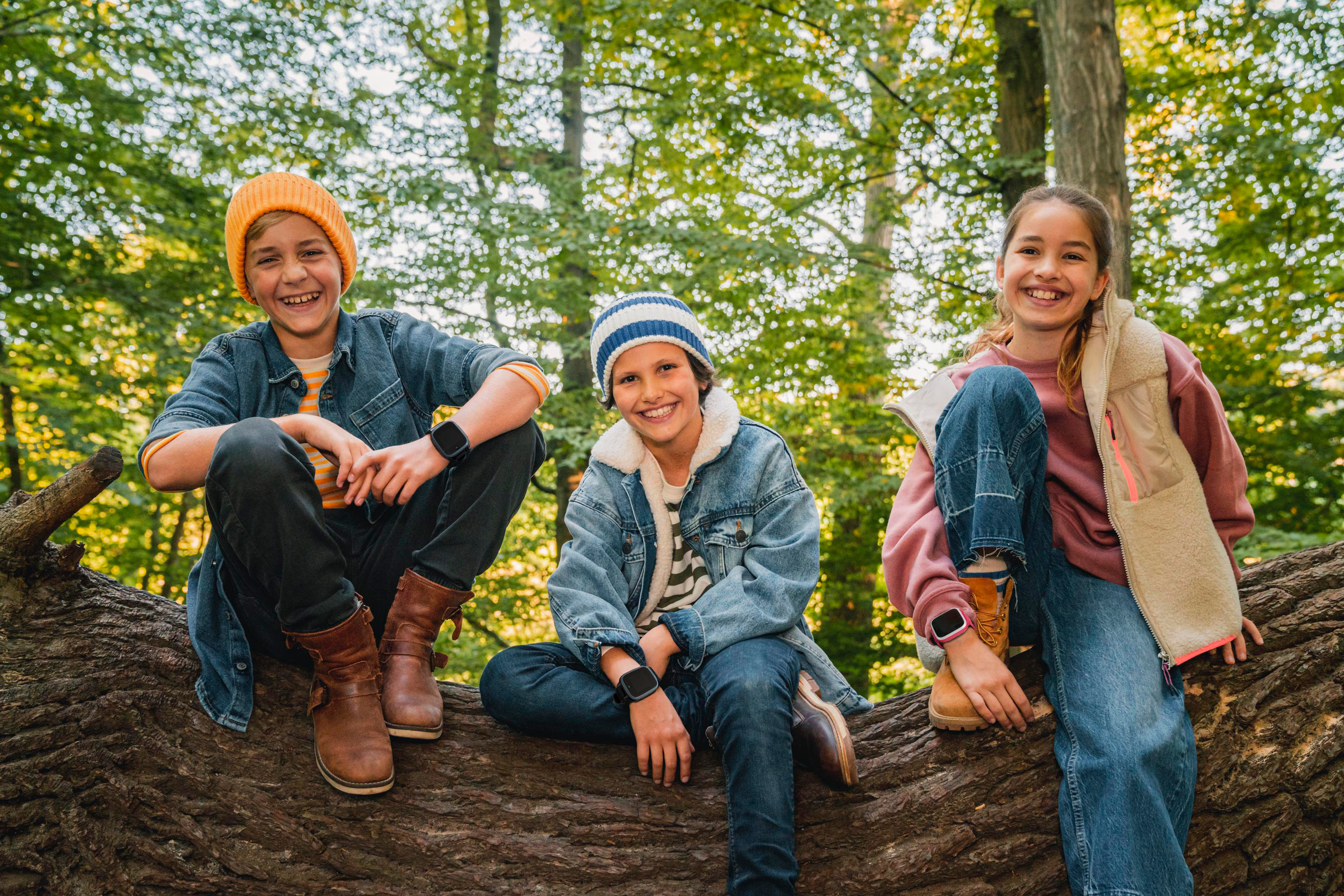 Drei Kinder sitzen auf einem Baumstamm im Wald, alle tragen Smartwatches und lächeln.