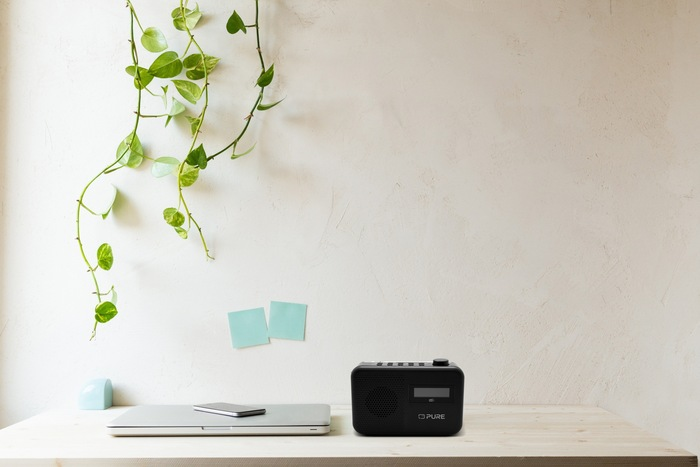 Un bureau avec un ordinateur portable, une radio et deux post-it. Des plantes vertes sont suspendues au-dessus.