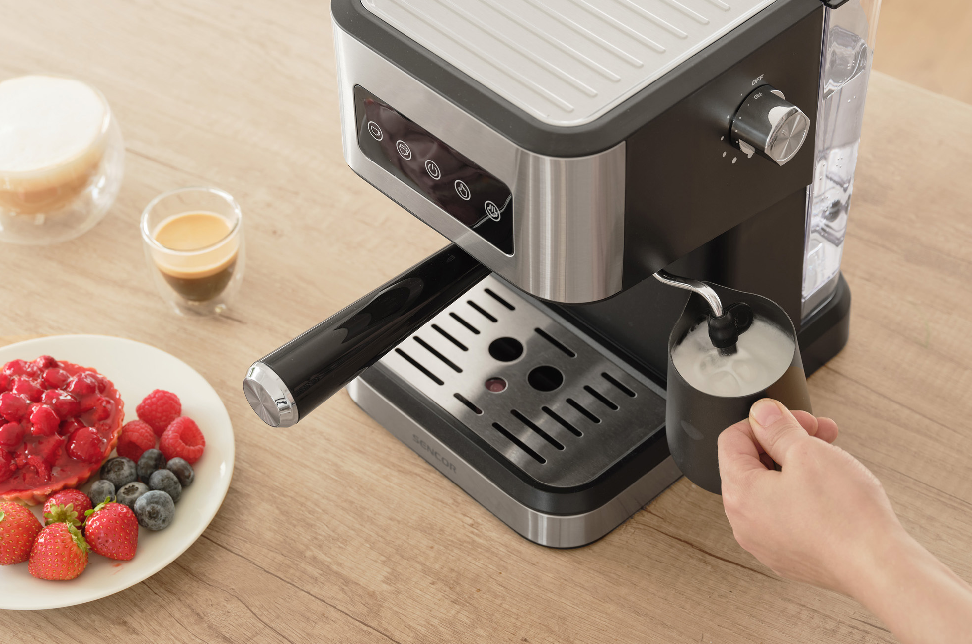 Esspresszó gép tejhabosítással, egy csésze eszpresszó és egy desszertes tányér a faasztalon.