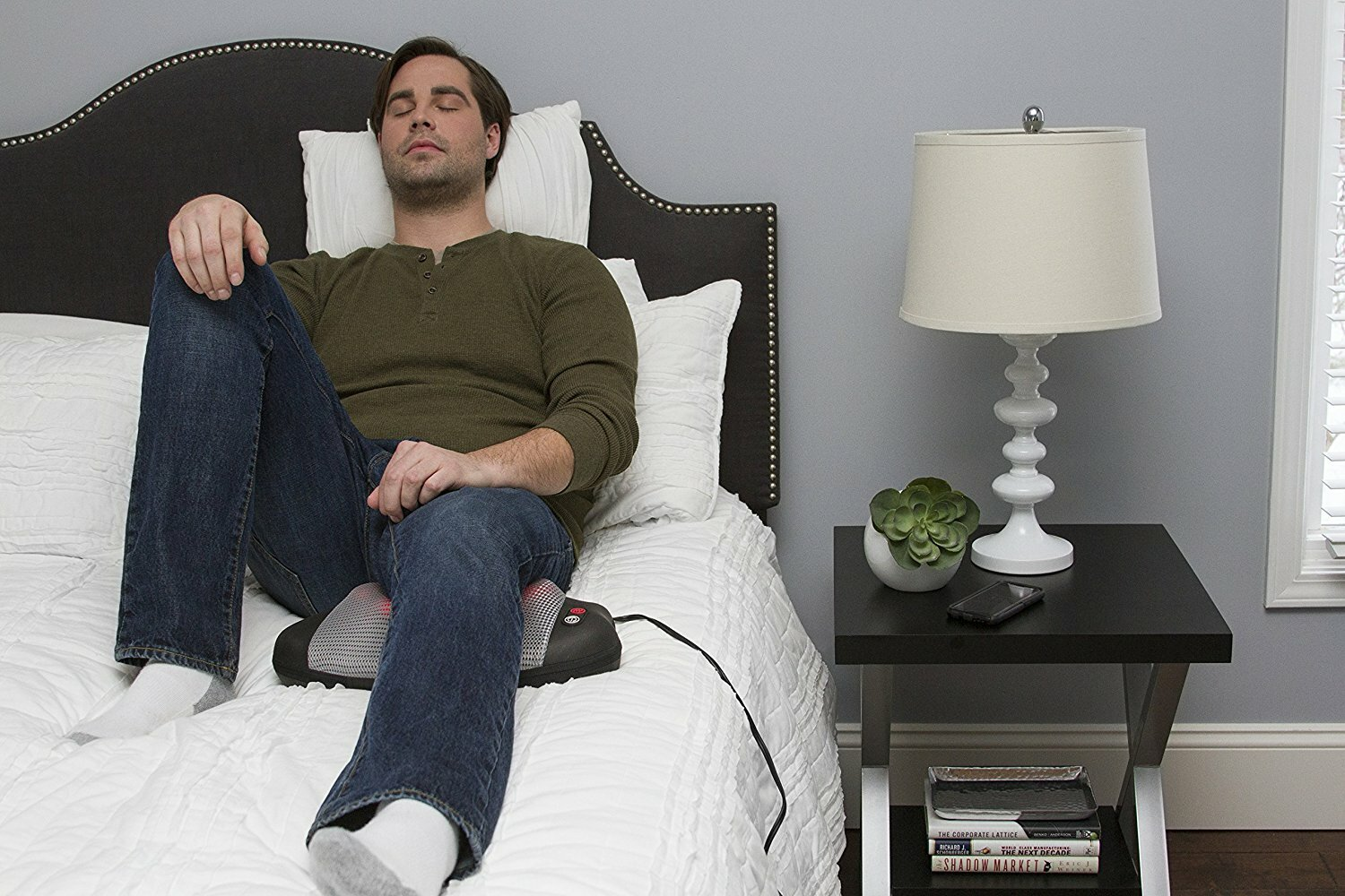 Mężczyzna w casualowych ubraniach odpoczywa na łóżku z masażerem nóg. Stolik nocny z lampą.