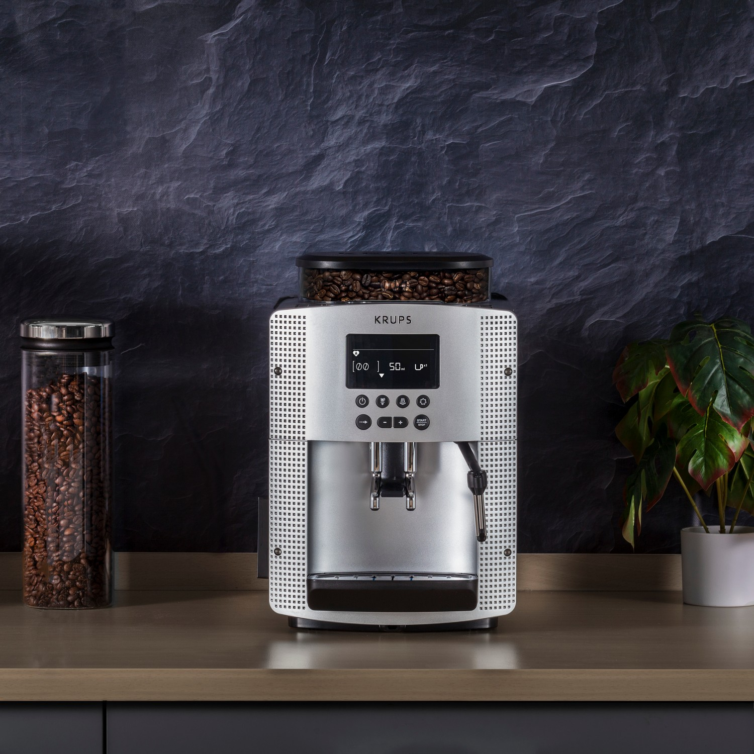 Silver Krups coffee machine with bean container. Coffee beans, plant and dark background.