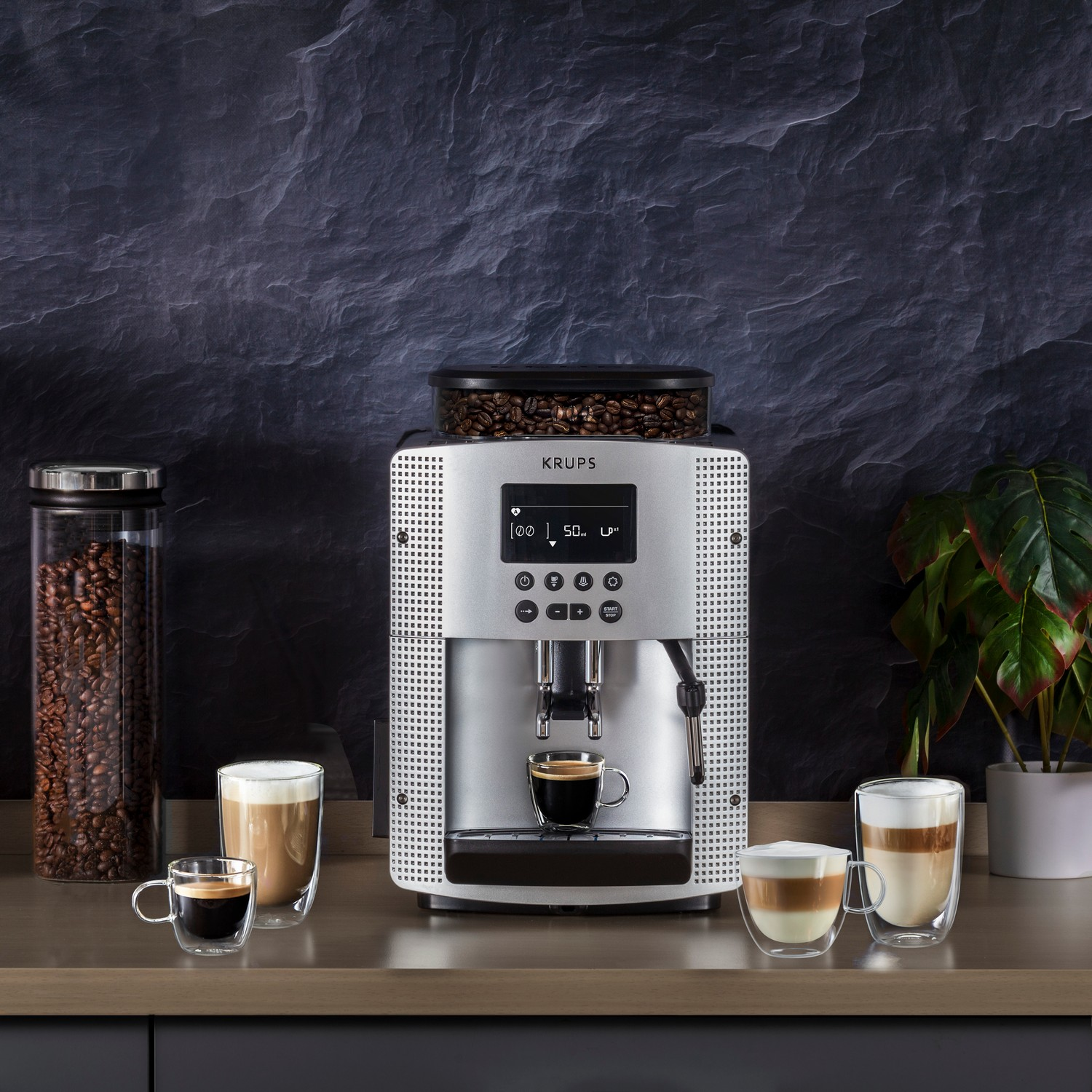 A silver Krups espresso machine with coffee cups, beans, and a plant on a counter.