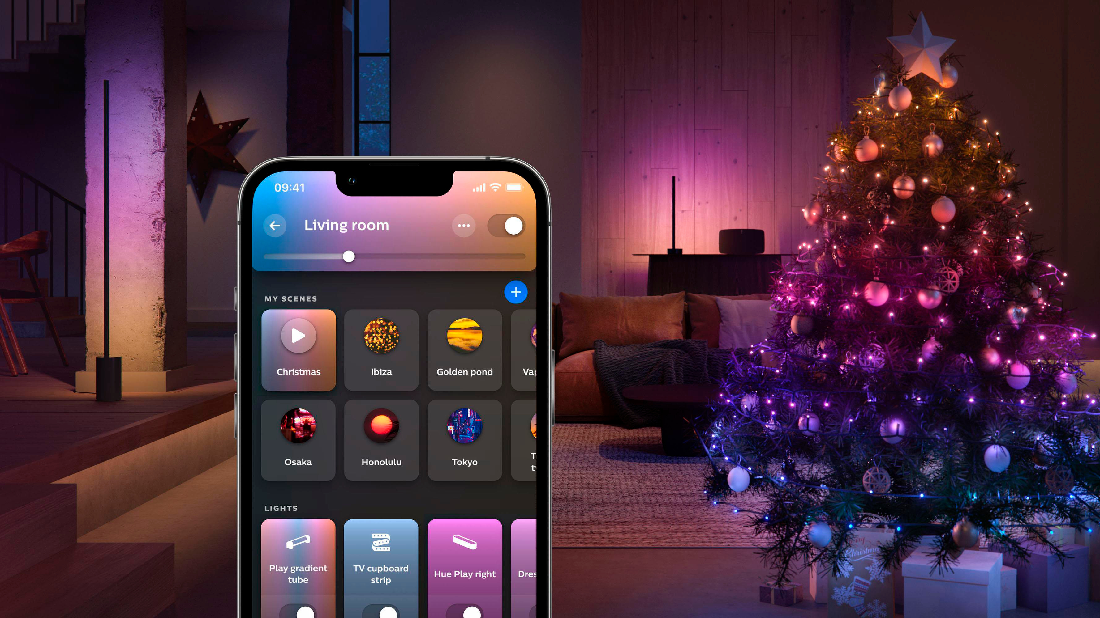 Ein Smartphone zeigt eine Smart-Home-App. Ein Weihnachtsbaum mit bunten Lichtern im Hintergrund.