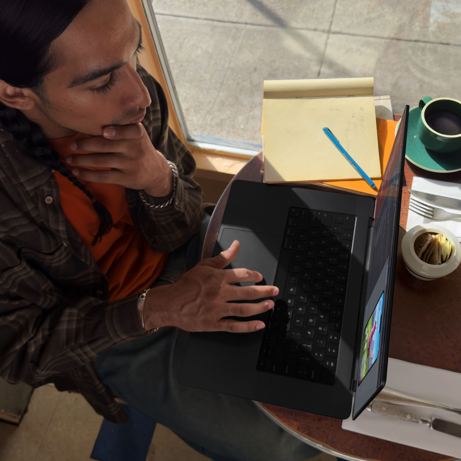 Un hombre usando una laptop negra en un café, con un cuaderno y una taza de café cerca.