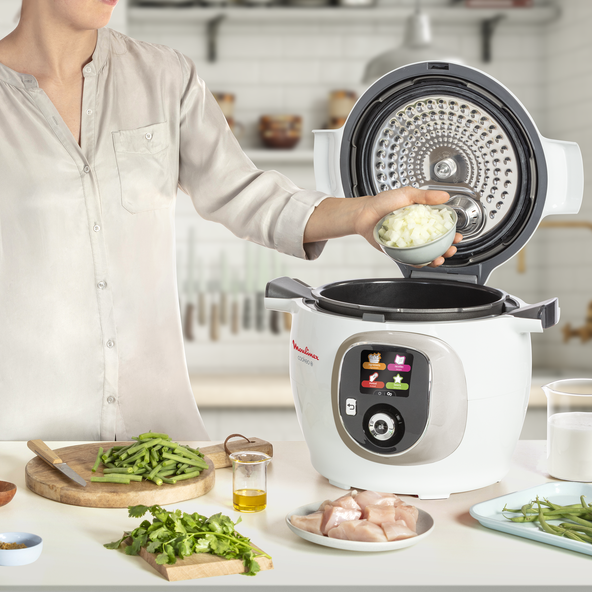 Femme ajoutant des oignons hachés à une cuisinière Moulinex blanche, ingrédients sur la table.