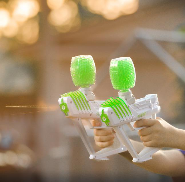 Zwei Wasserspielzeugpistolen schießen grüne Gelbälle. Unscharfer Hintergrund. Hände halten Pistolen.