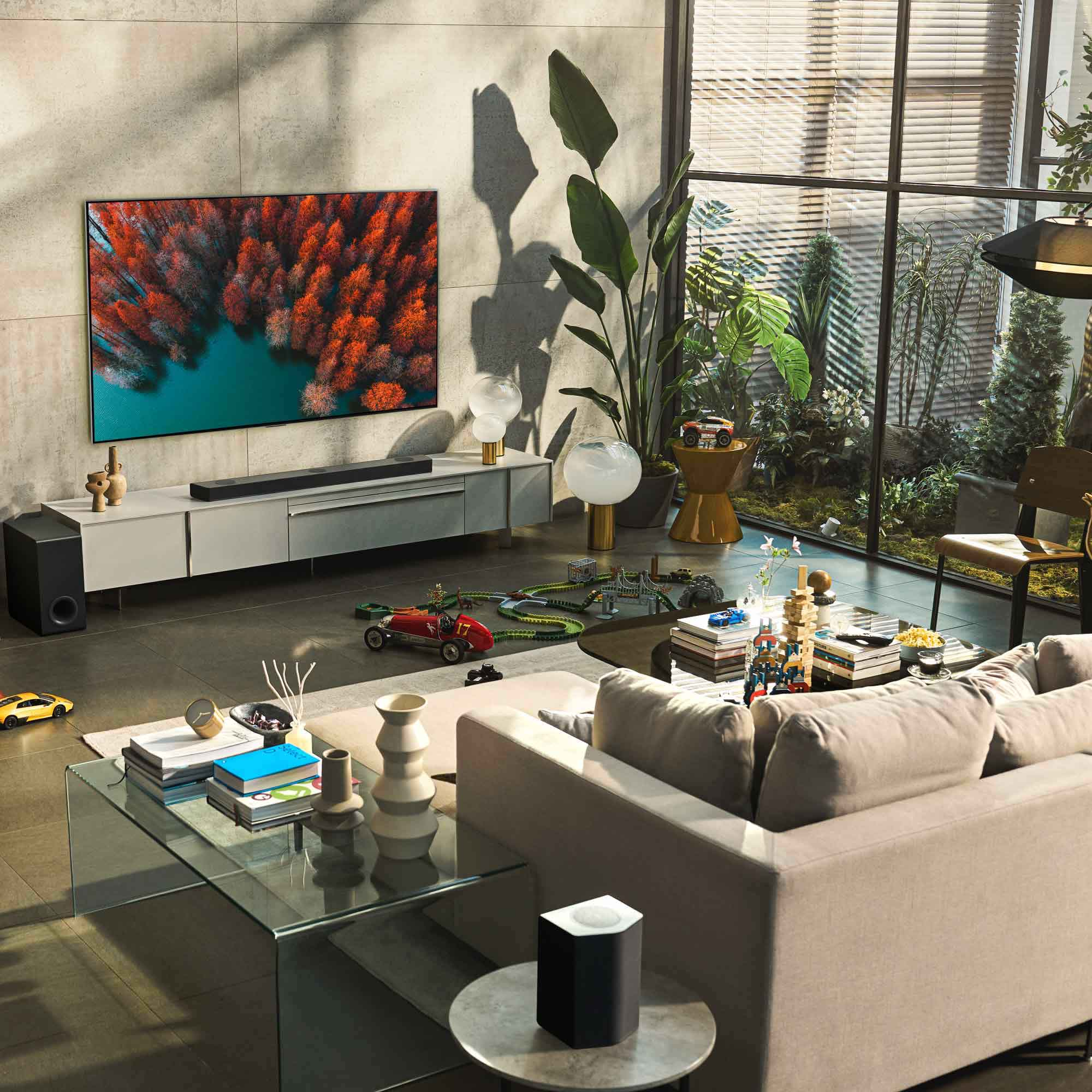 Escena de salón con un televisor grande, barra de sonido y sofá. Hay plantas y decoración.