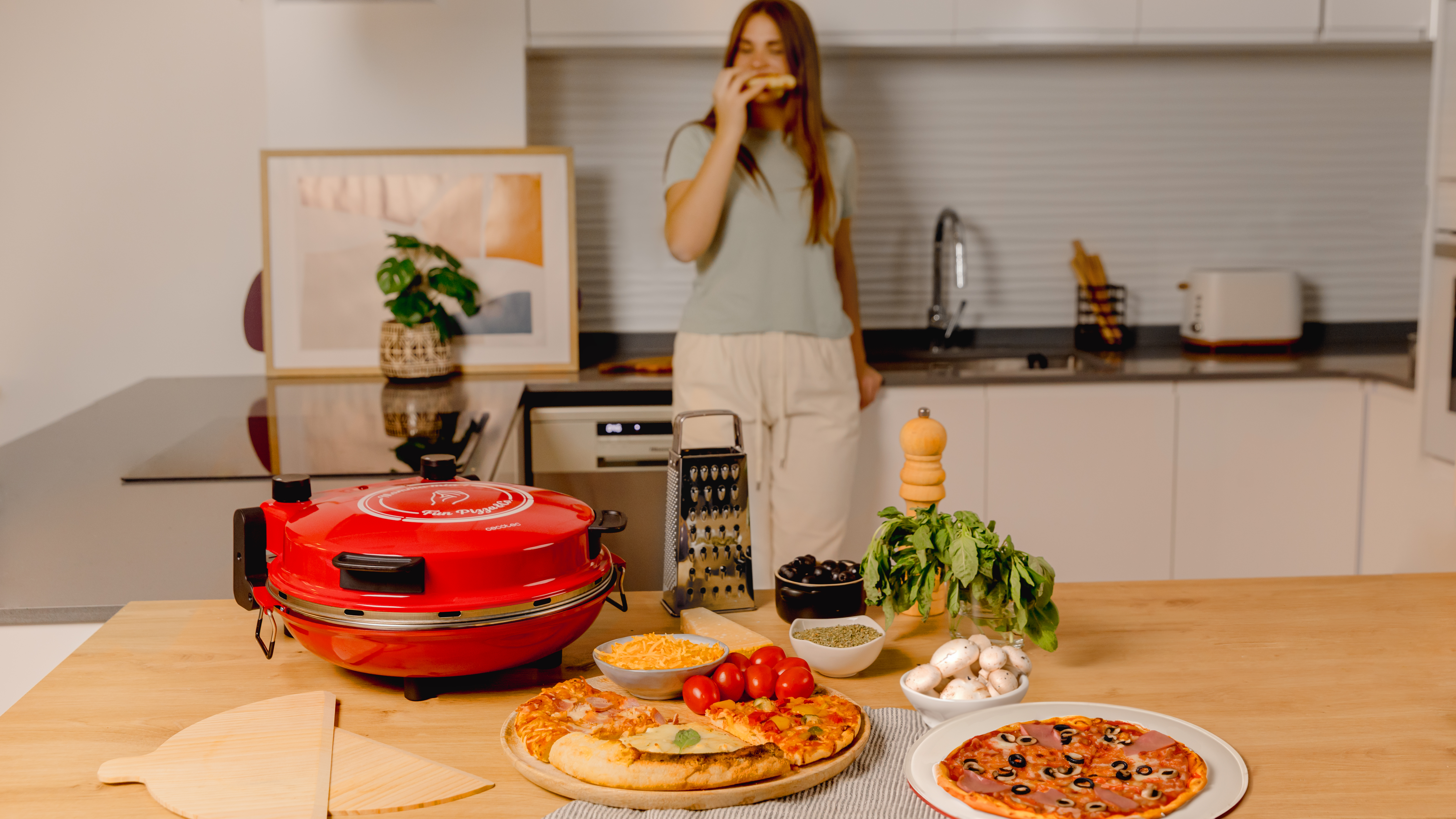 Una mujer comiendo pizza cerca de un horno de pizza, pizza e ingredientes en una mesa.