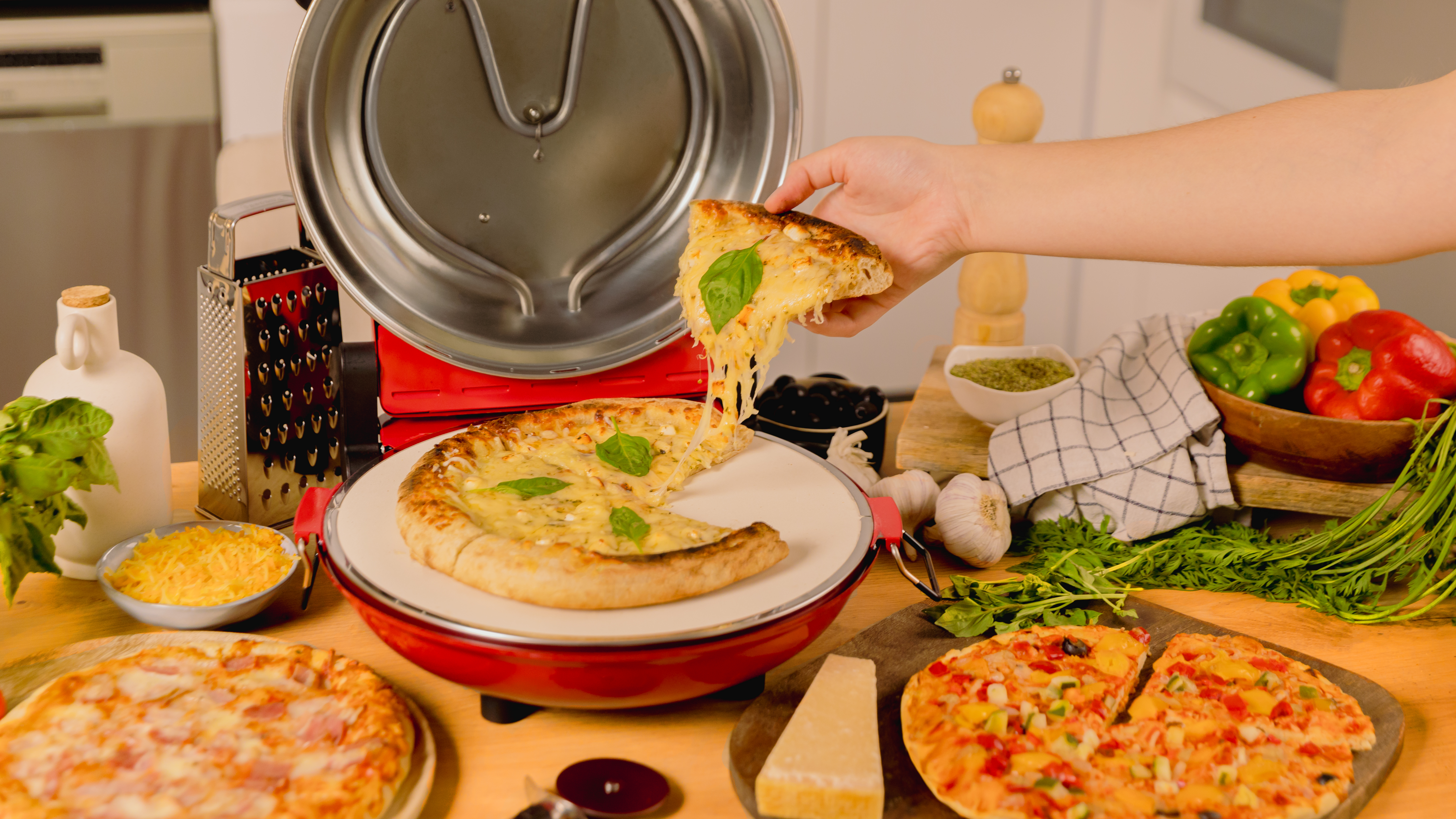 Una mano sostiene una porción de pizza sobre una pizza. Ingredientes y otras pizzas en la mesa.