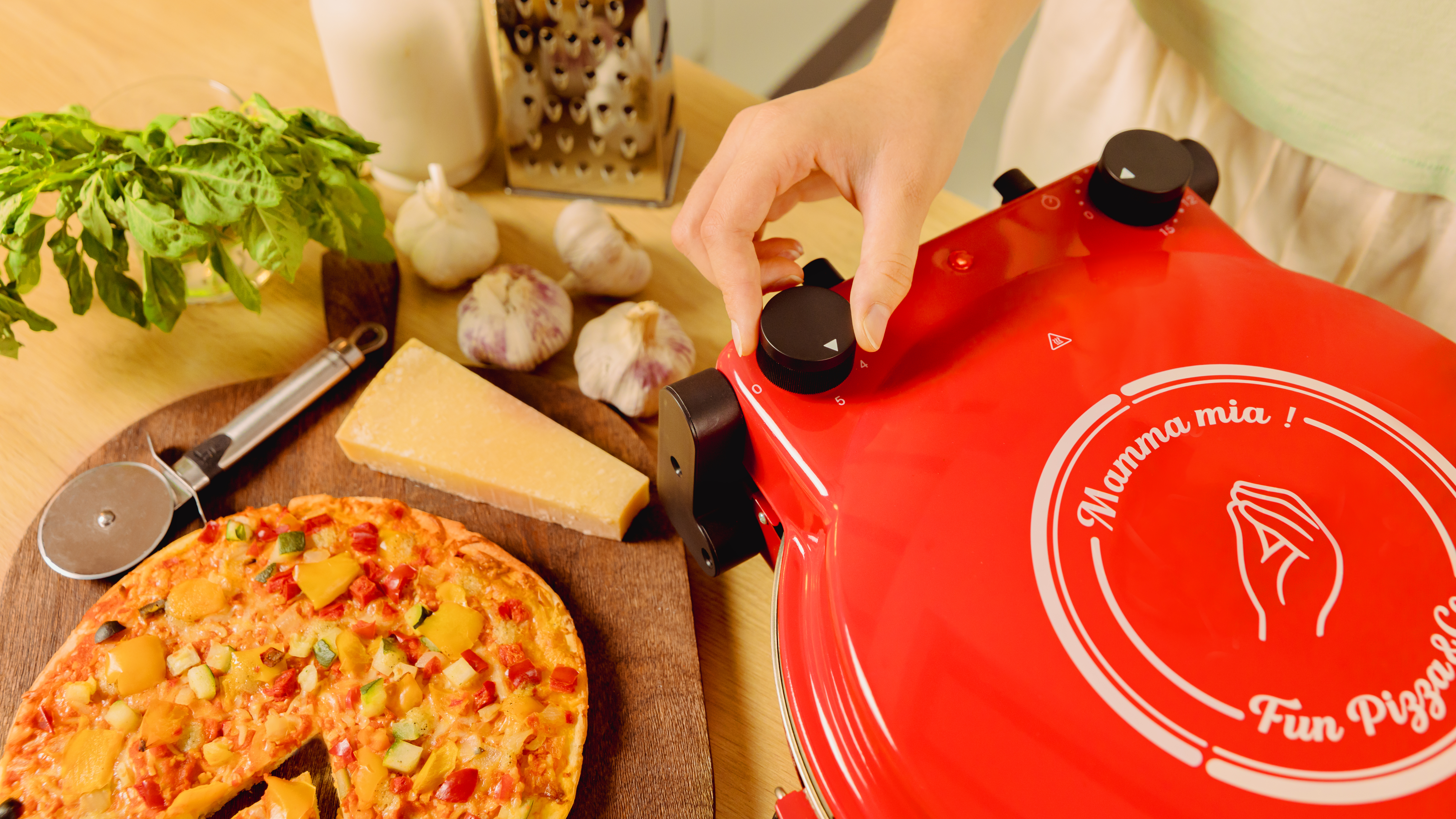Una mano ajusta una perilla de horno de pizza roja. Pizza, ingredientes y un cortador en la mesa.