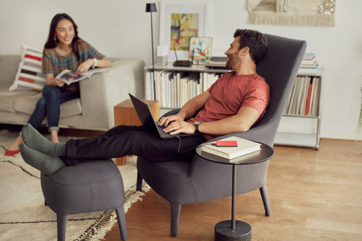 Mężczyzna z laptopem w fotelu, druga osoba czytająca na kanapie w tle.