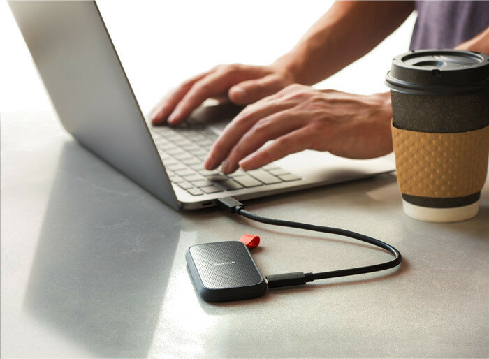 Osoba korzystająca z laptopa z podłączonym czarnym przenośnym dyskiem SSD. Po prawej stronie kubek kawy.