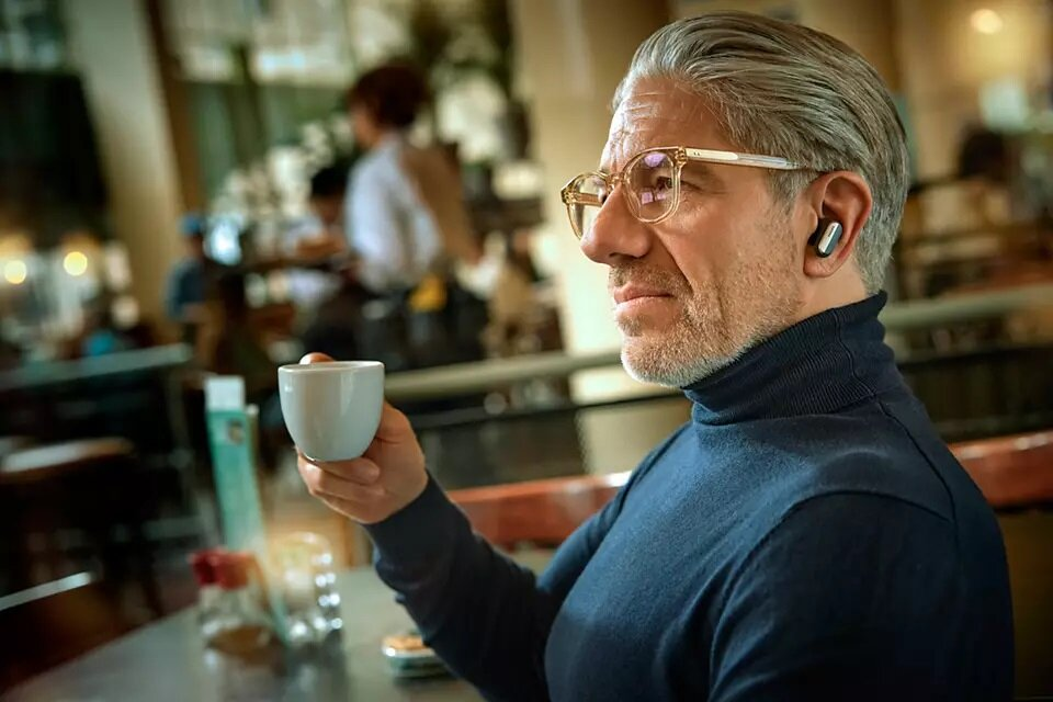Mężczyzna w kawiarni trzymający filiżankę, w okularach i słuchawkach.