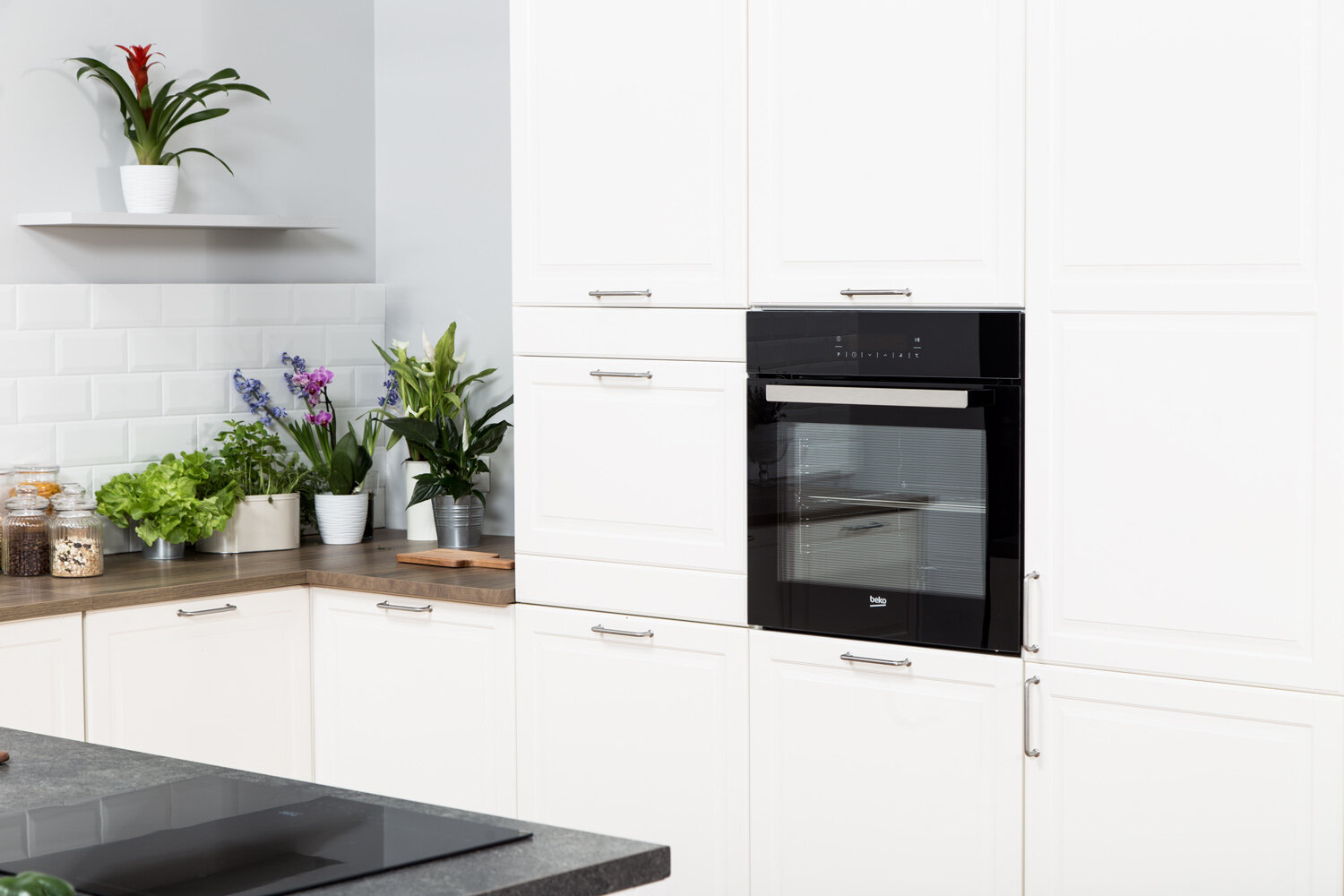 Nowoczesna biała kuchnia z wbudowanym piekarnikiem i szafkami. Widoczne są również rośliny i blat.