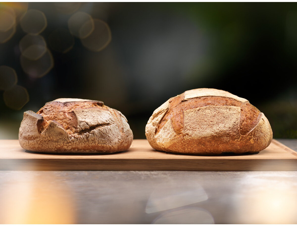 Dwa bochenki rustykalnego chleba na drewnianej desce. Chleb jest złocisty z teksturowaną skórką. Tło jest rozmyte.