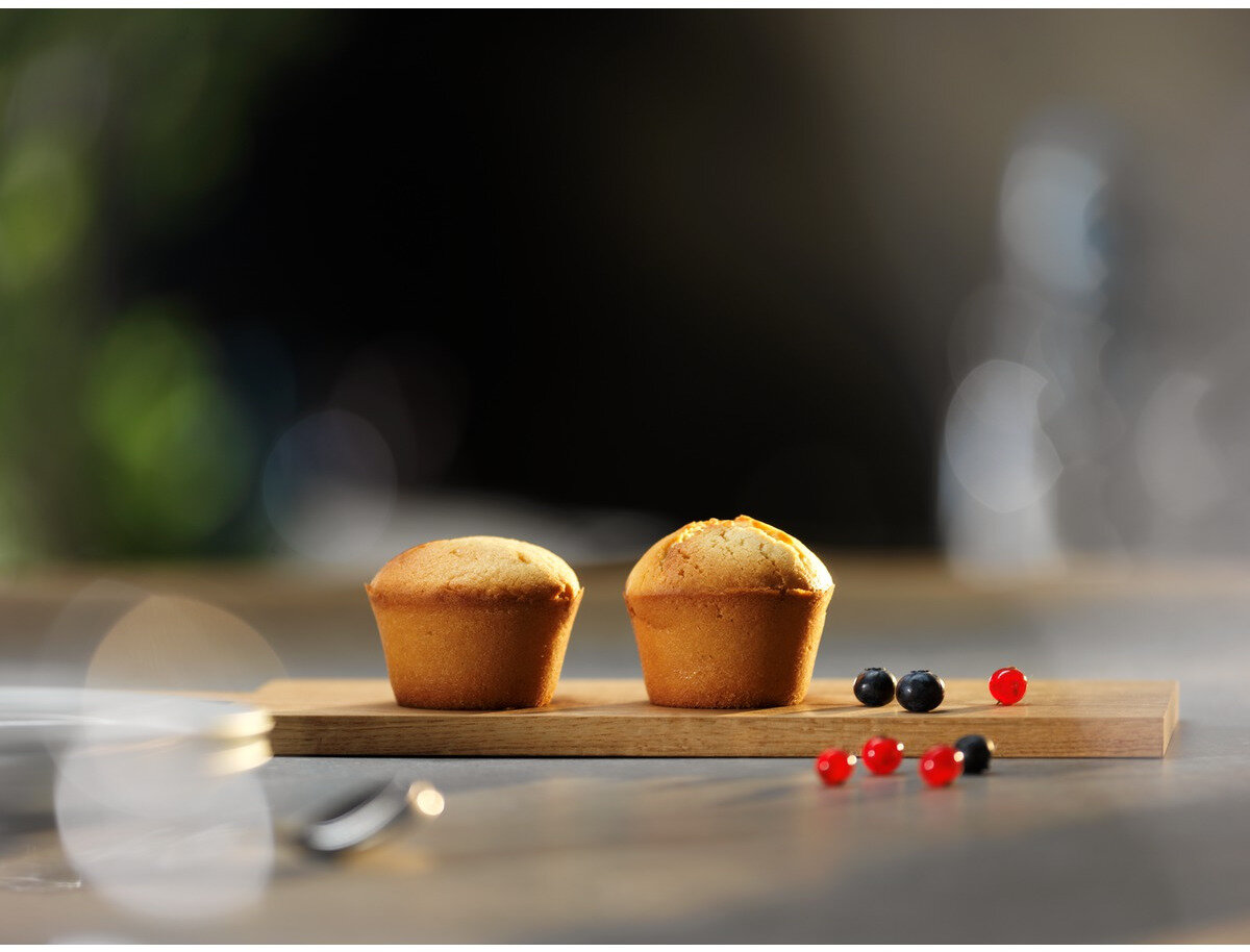Dwa muffiny na drewnianej desce z jagodami. Kilka jagód jest również rozrzuconych po stole. Tło jest rozmyte.