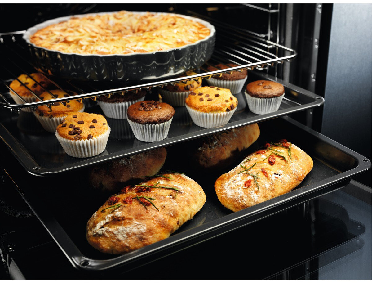 Piekarnik z trzema tacami z wypiekami: ciastem, babeczkami i chlebem. Chleb ma zioła i pomidory. Piekarnik jest czarny.