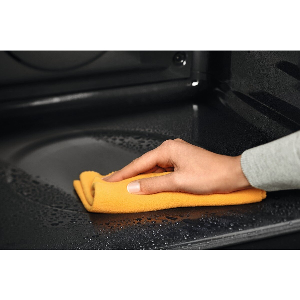 Ręka wyciera żółtą szmatką mokrą, czarną powierzchnię piekarnika.