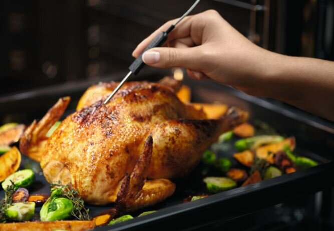 Osoba wkładająca termometr do mięsa do pieczonego kurczaka w patelni z warzywami.