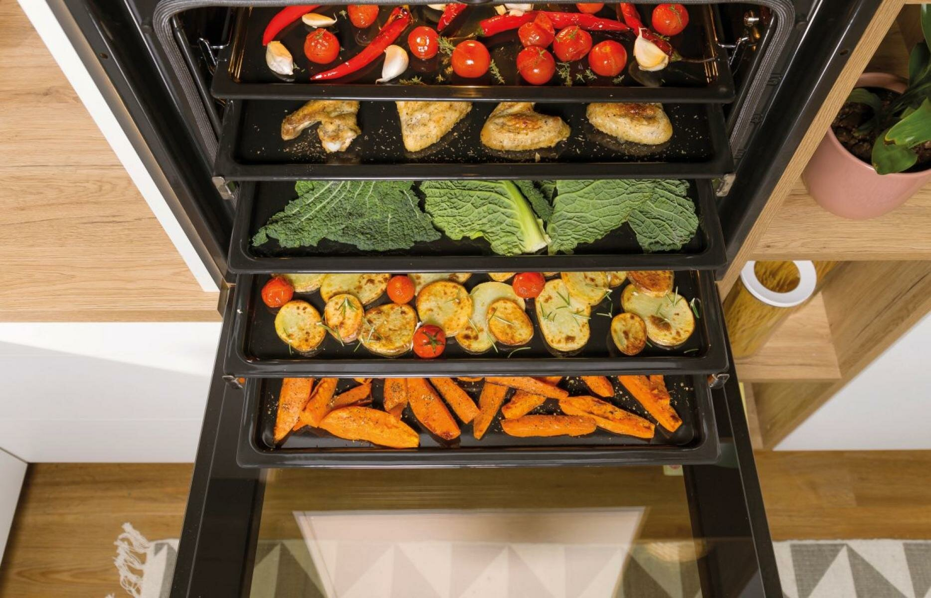 Piekarnik z tacami jedzenia: pomidory, papryka, kurczak, kapusta, ziemniaki i marchew. Drewniana półka i biała szafka obok.