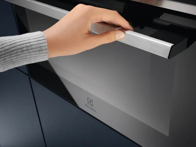 Dłoń otwierająca drzwi piekarnika, srebrny uchwyt, marka Electrolux, niebieskie tło kuchni.