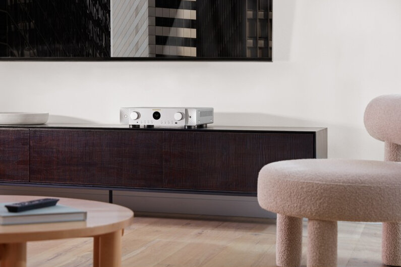 Srebrny odbiornik stereo na ciemnobrązowej szafce. Okrągły stół i krzesło są na pierwszym planie.