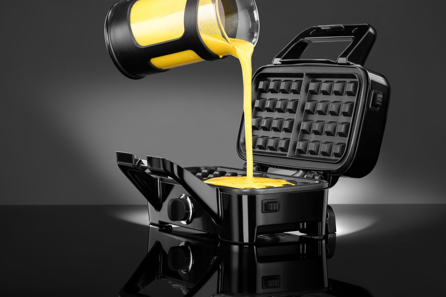 Ciasto na gofry wylewane z czarnego dozownika do czarnej gofrownicy. Tło jest ciemne.