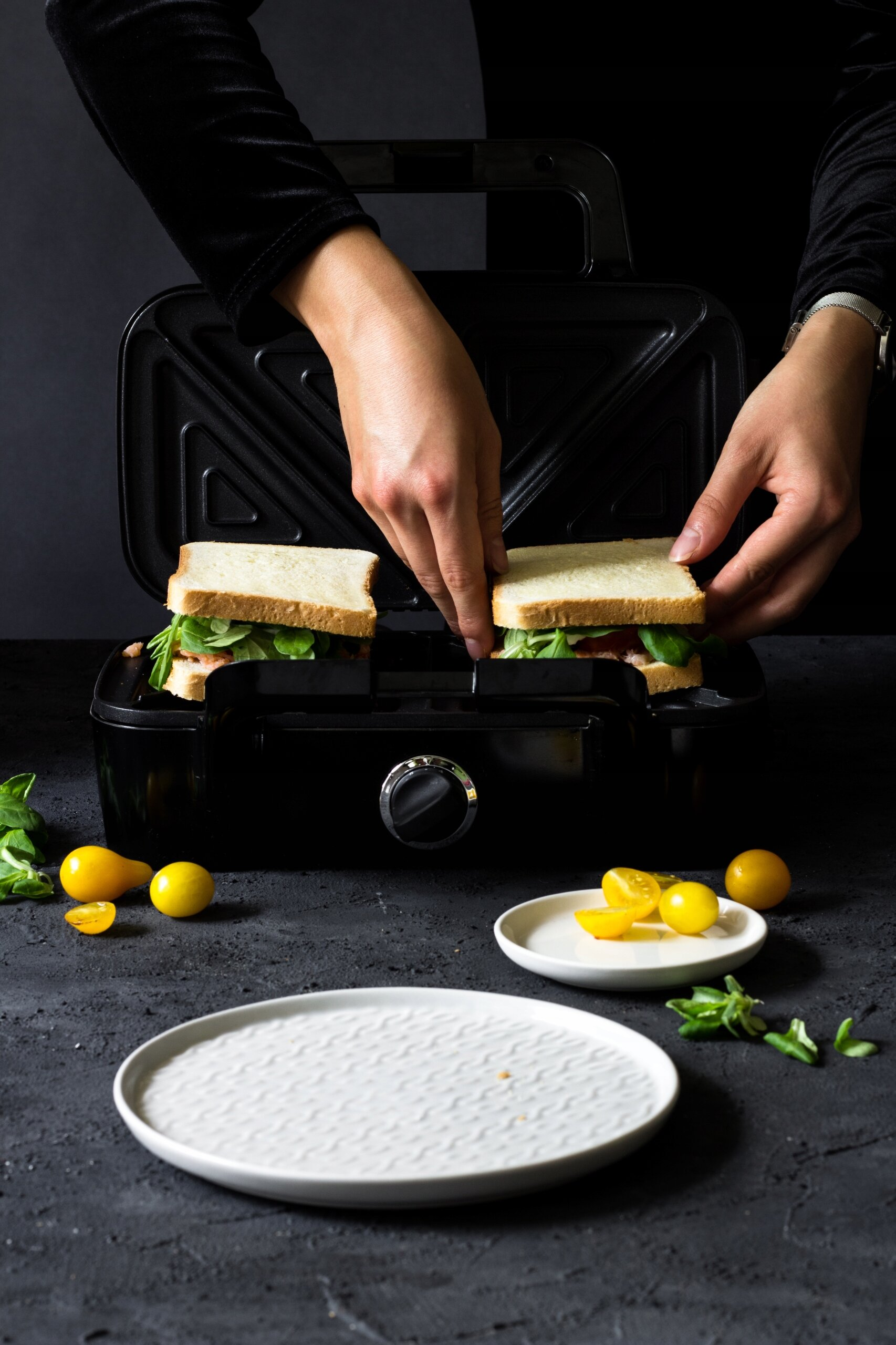 Ręce wkładające połówki kanapek do czarnego opiekacza. Pomidory i talerz.