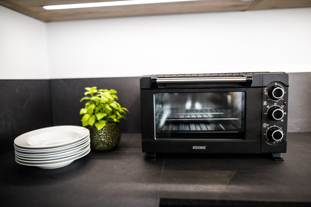 Czarny piekarnik stoi na blacie. Ułożone białe talerze i roślina po lewej. Piekarnik ma pokrętła i szklane drzwi.