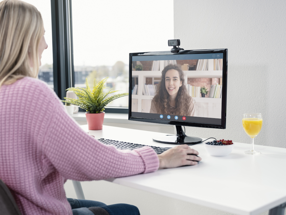 Femme en appel vidéo. Moniteur montre une autre femme souriante. Plante, fruits et verre à côté.