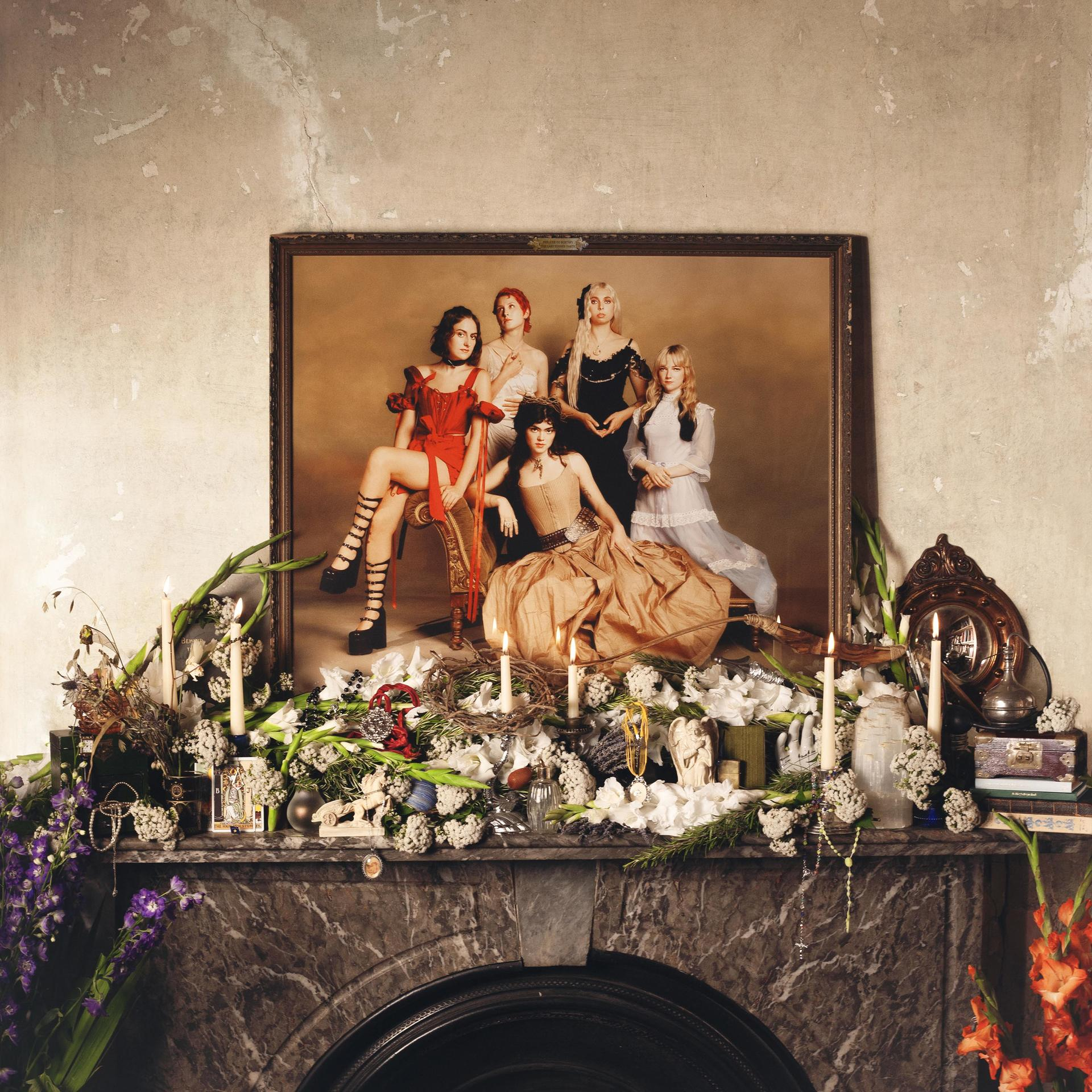 Een ingelijst schilderij van zes vrouwen, kaarsen en bloemen op een schoorsteenmantel. Interieur, warme tonen.