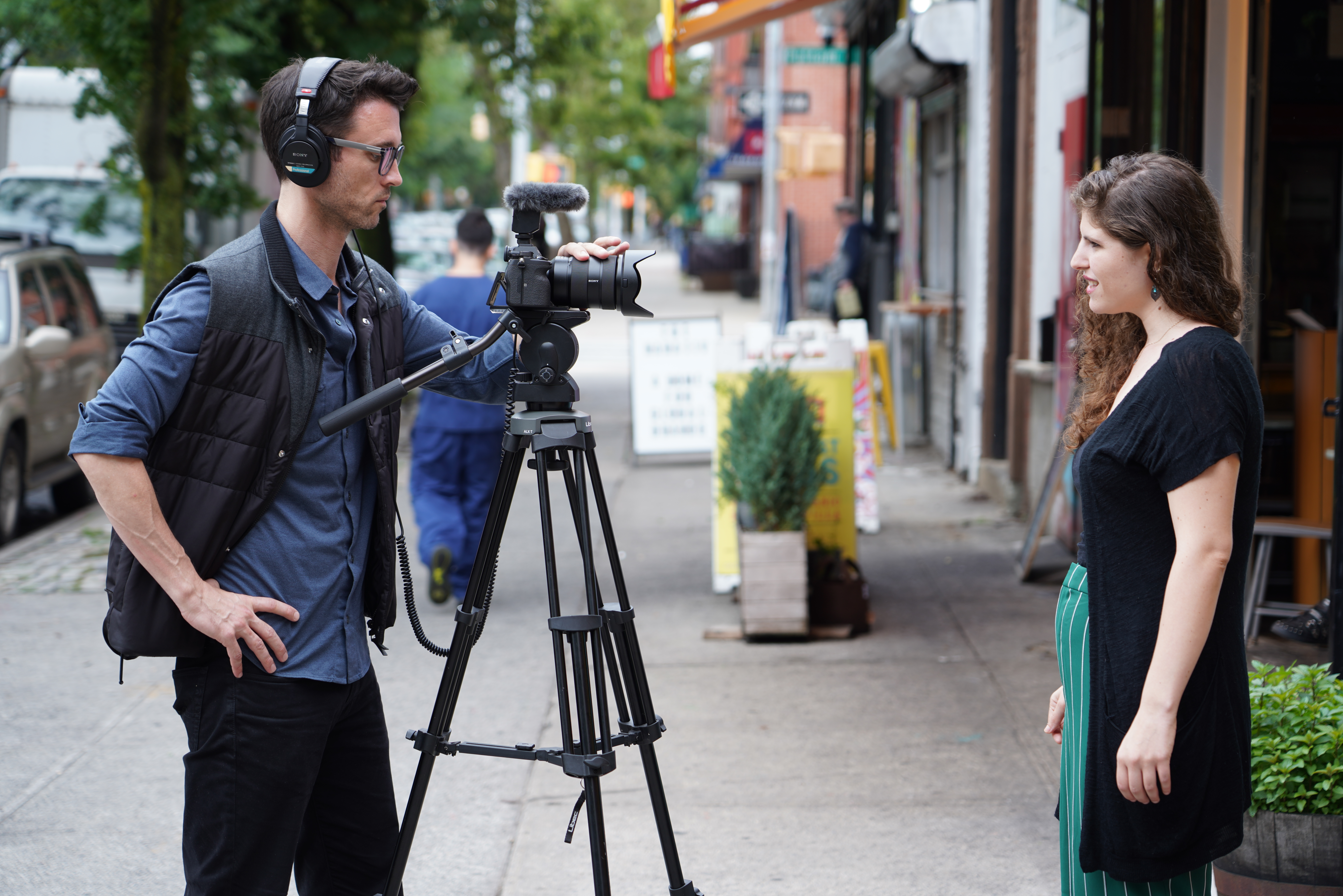 Scène de tournage : Un homme avec une caméra sur trépied interviewe une femme sur un trottoir. Fond de ville.