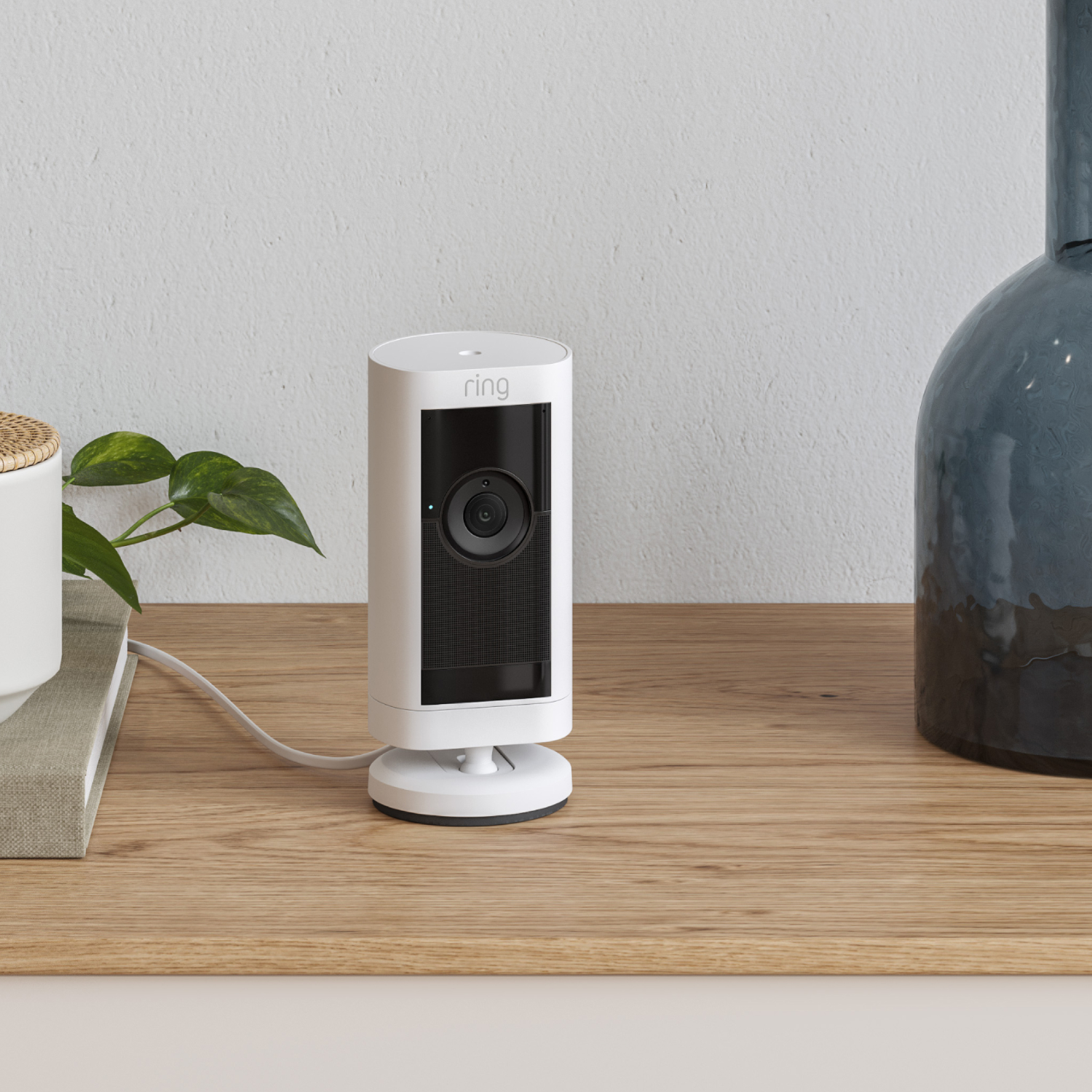 White Ring security camera on a wooden table. A plant and a blue vase are in the background.