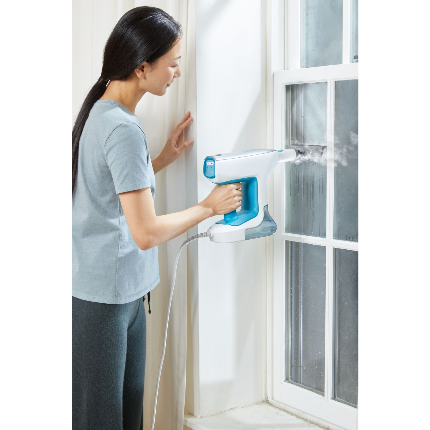A woman using a hand-held steam cleaner on a window, white wall.