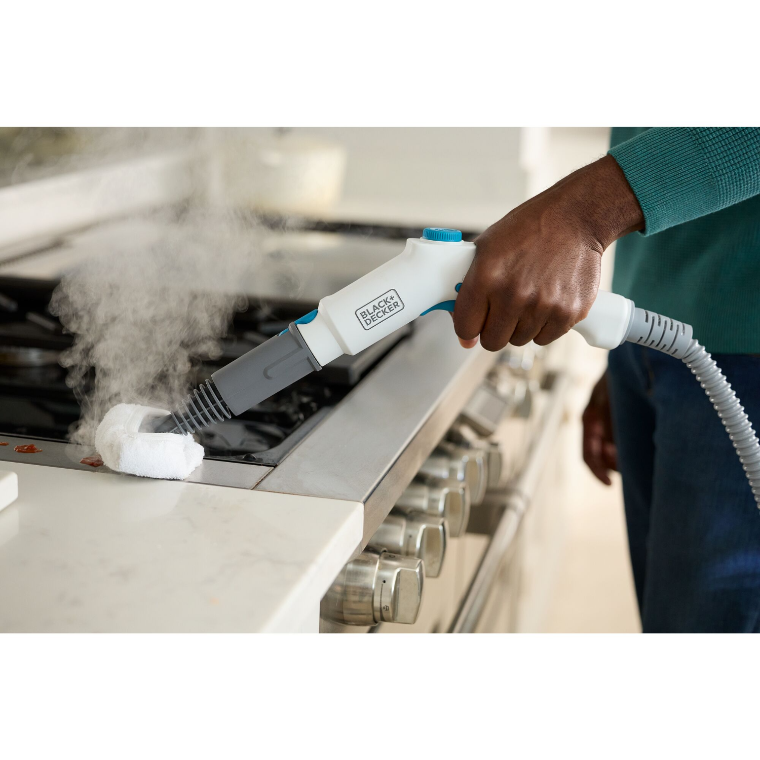 A person cleaning a stainless steel stove with a black and white steam cleaner. The background is a kitchen.