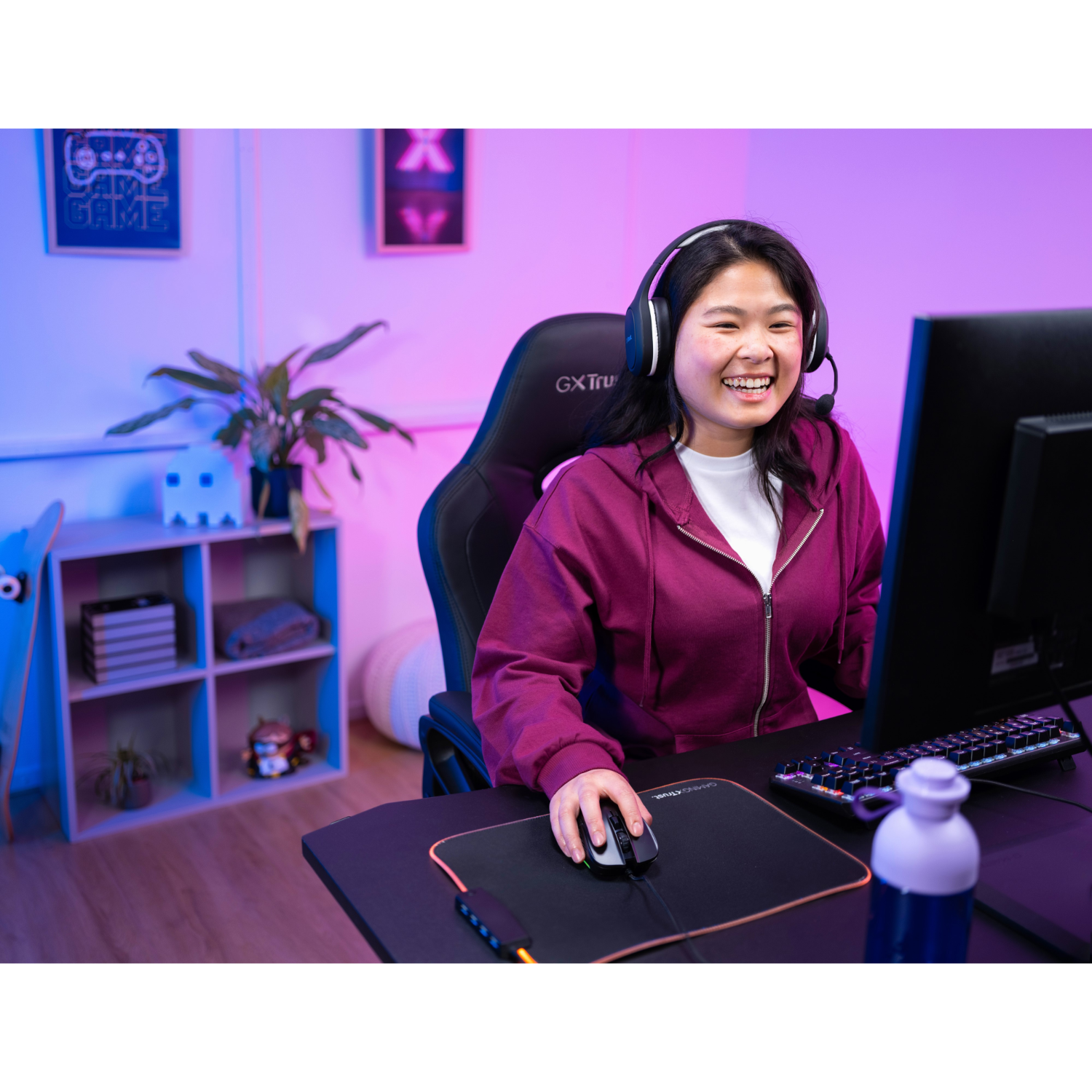 A young woman smiles while gaming on a computer, wearing a headset and using a mouse.