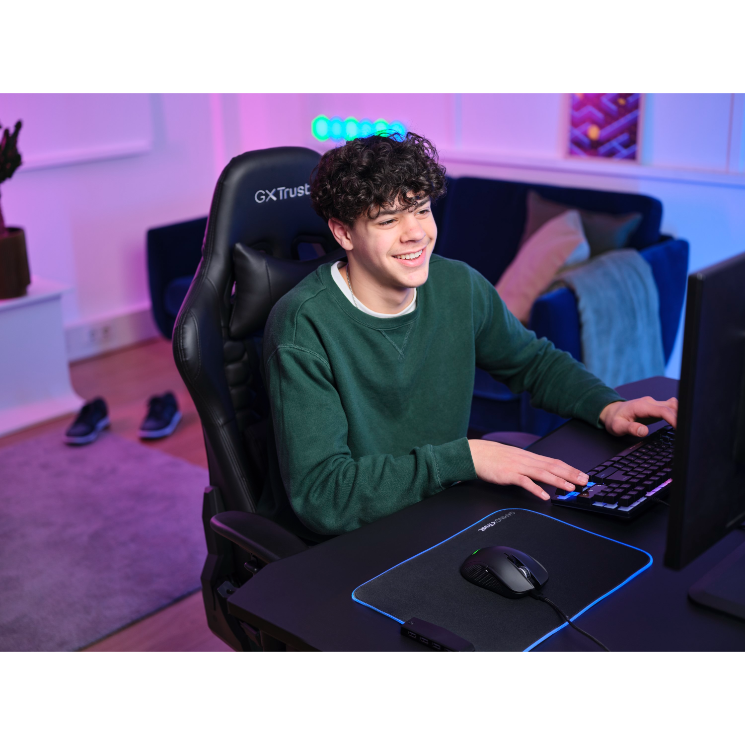 Young man sitting at a desk, smiling while gaming. He has a keyboard, mouse, and monitor. The background is colorful.