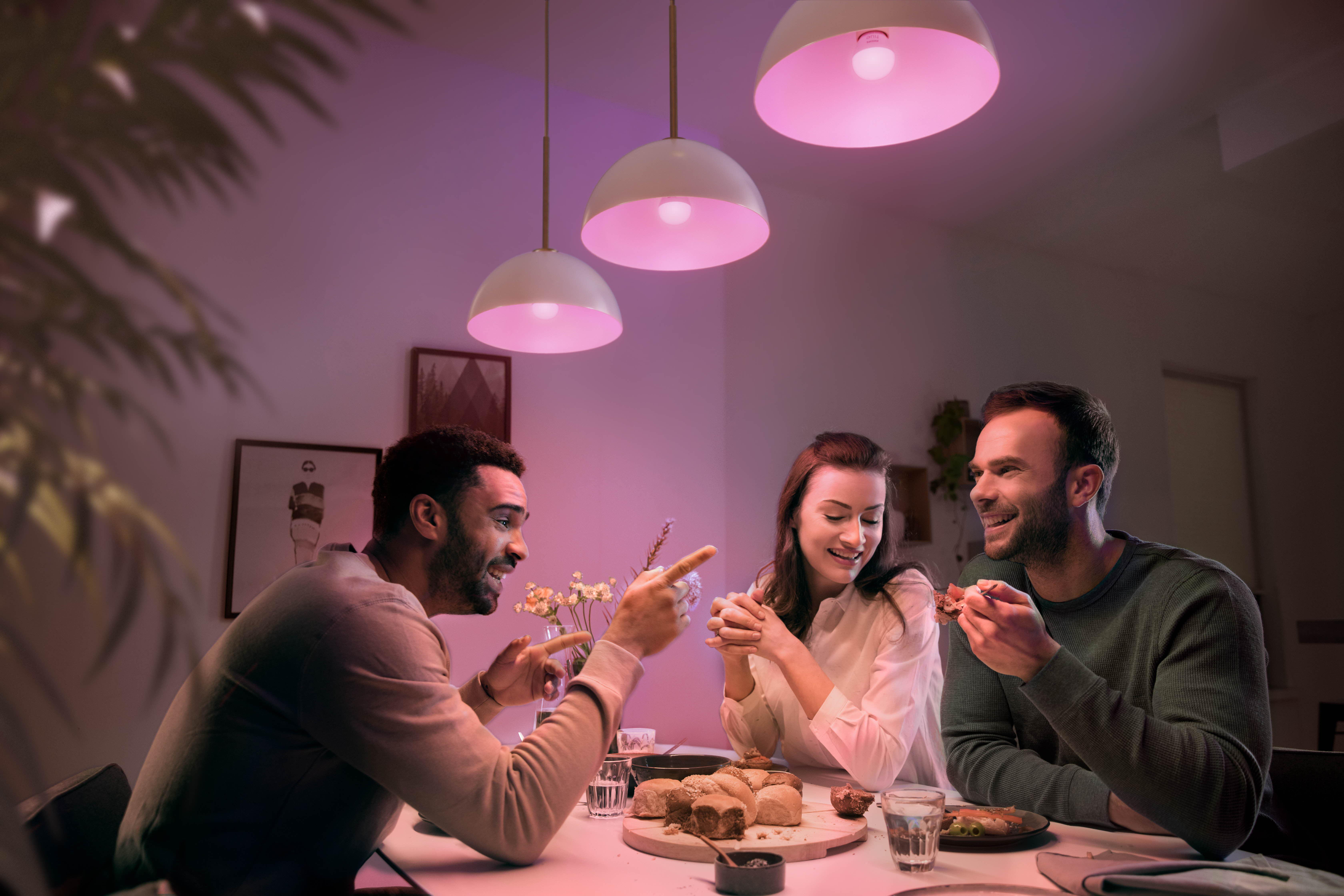 Drei Personen an einem Tisch, beleuchtet von rosa Lichtern, mit Essen und Getränken.