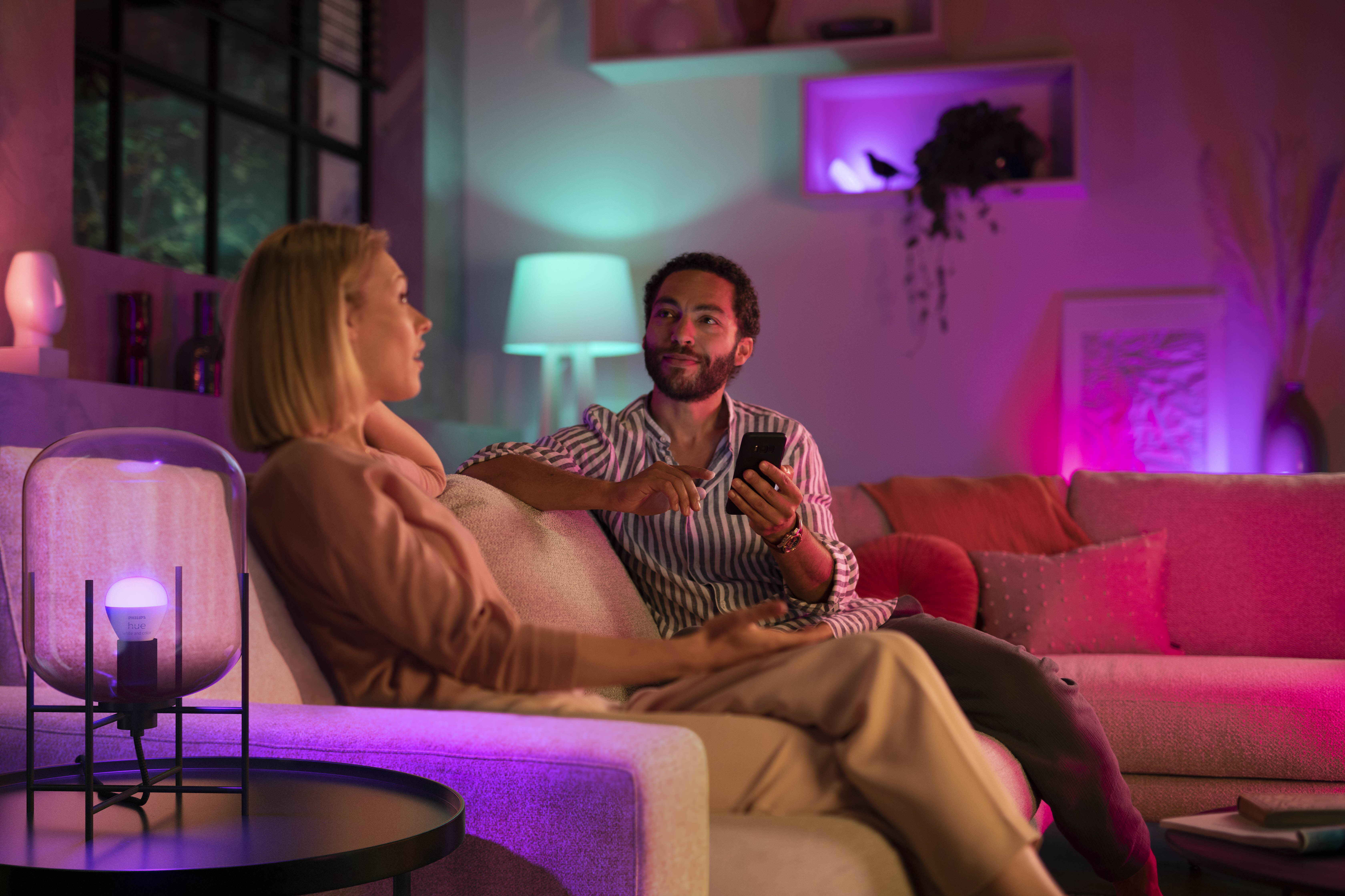 Zwei Personen auf einem Sofa mit bunten Lichtern um sie herum. Eine Lampe steht links.