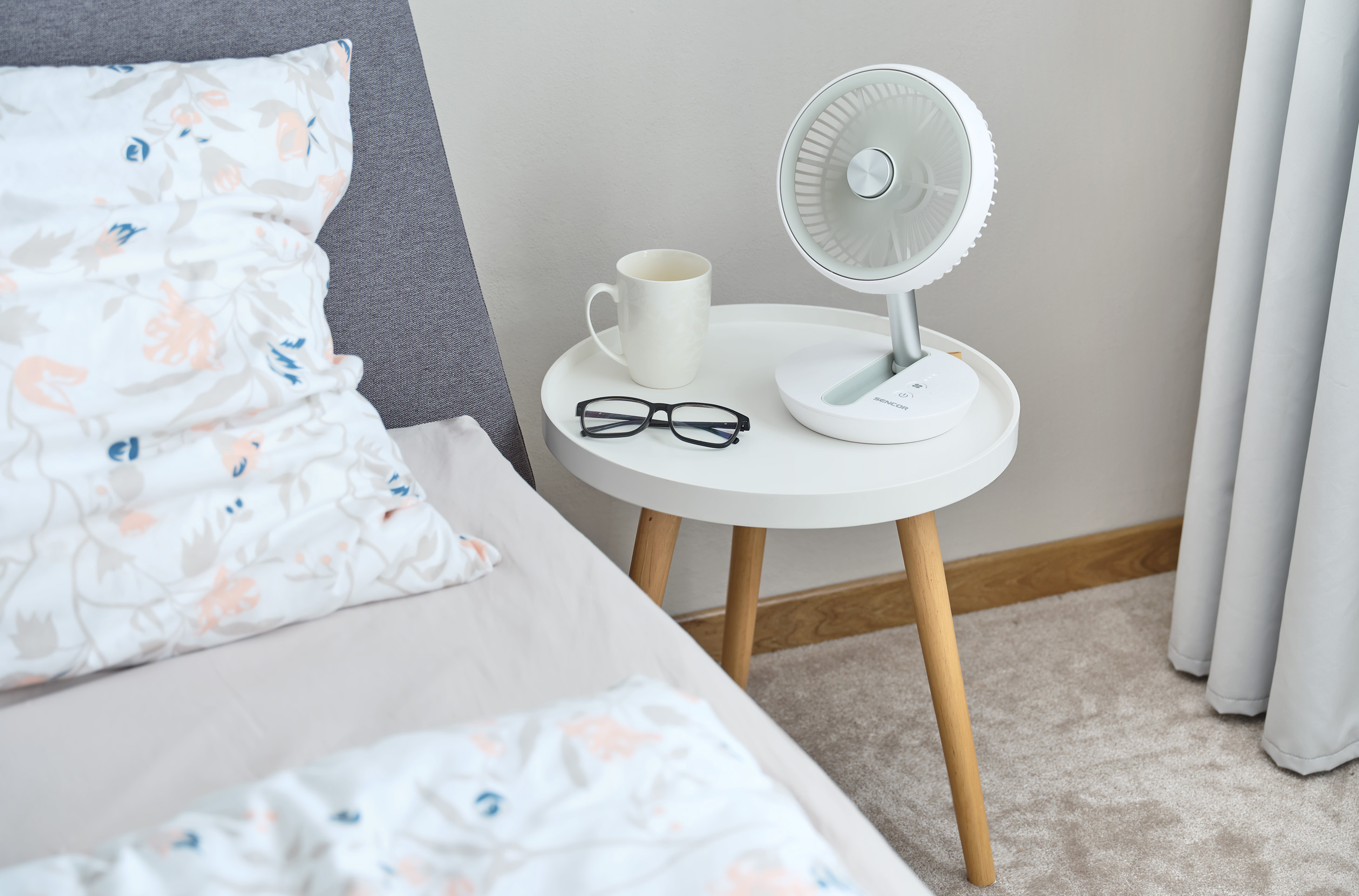 Fehér ventilátor az asztalon, bögre, szemüveg, ágy virágos ágyneművel.