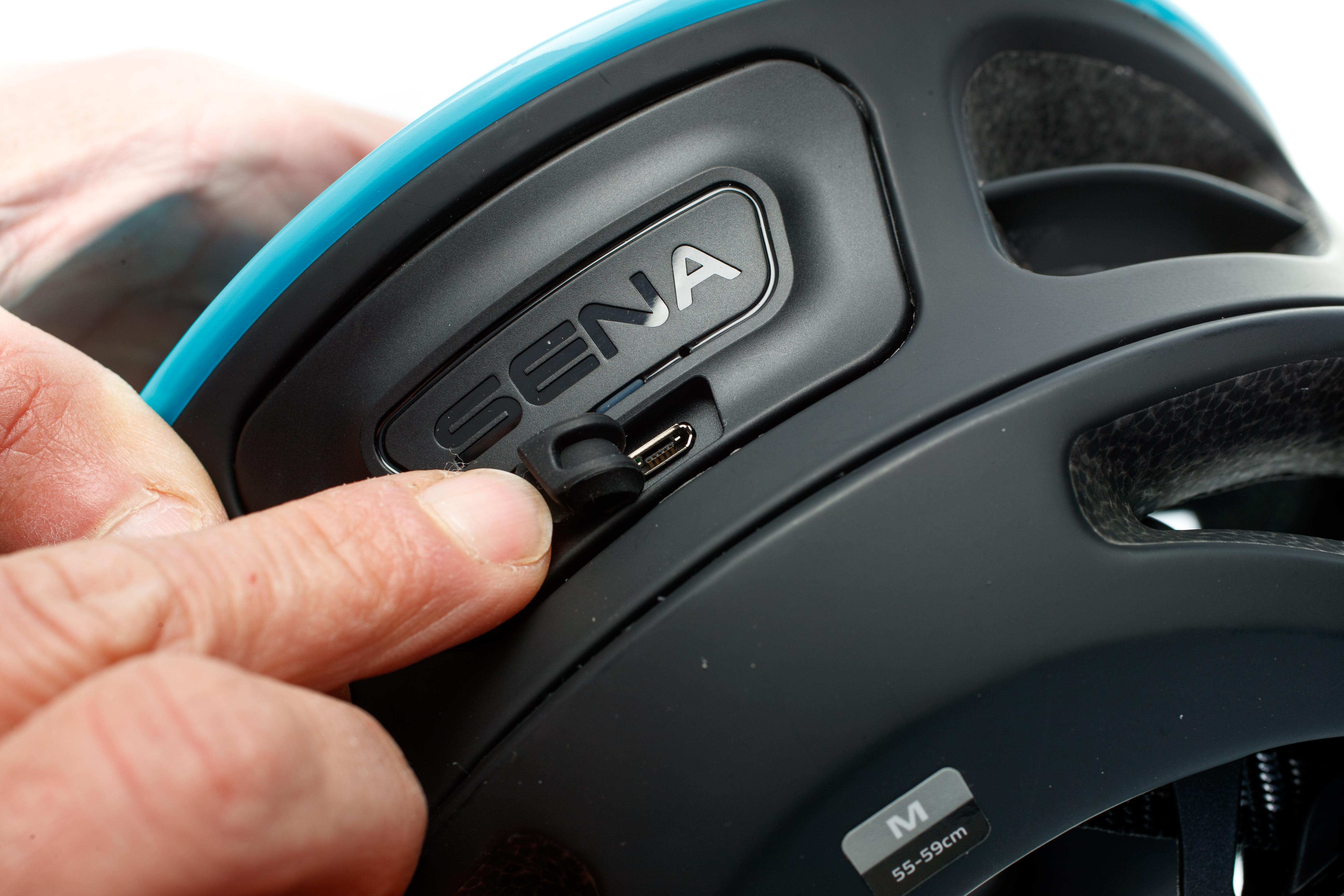 Nahaufnahme eines schwarzen und blaugrünen Fahrradhelms mit der Aufschrift 'SENA'. Ein Finger zeigt auf einen USB-Anschluss.