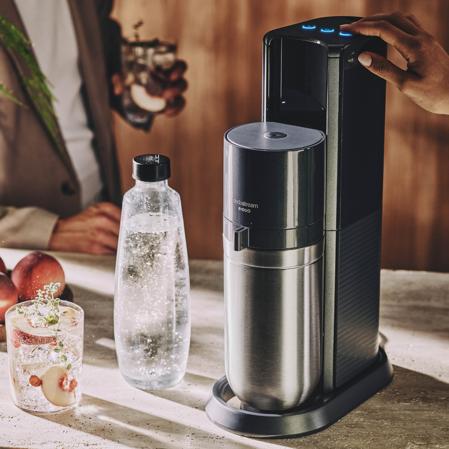 SodaStream-machine met een fles en een glas frisdrank met fruit. Een hand drukt op een knop.