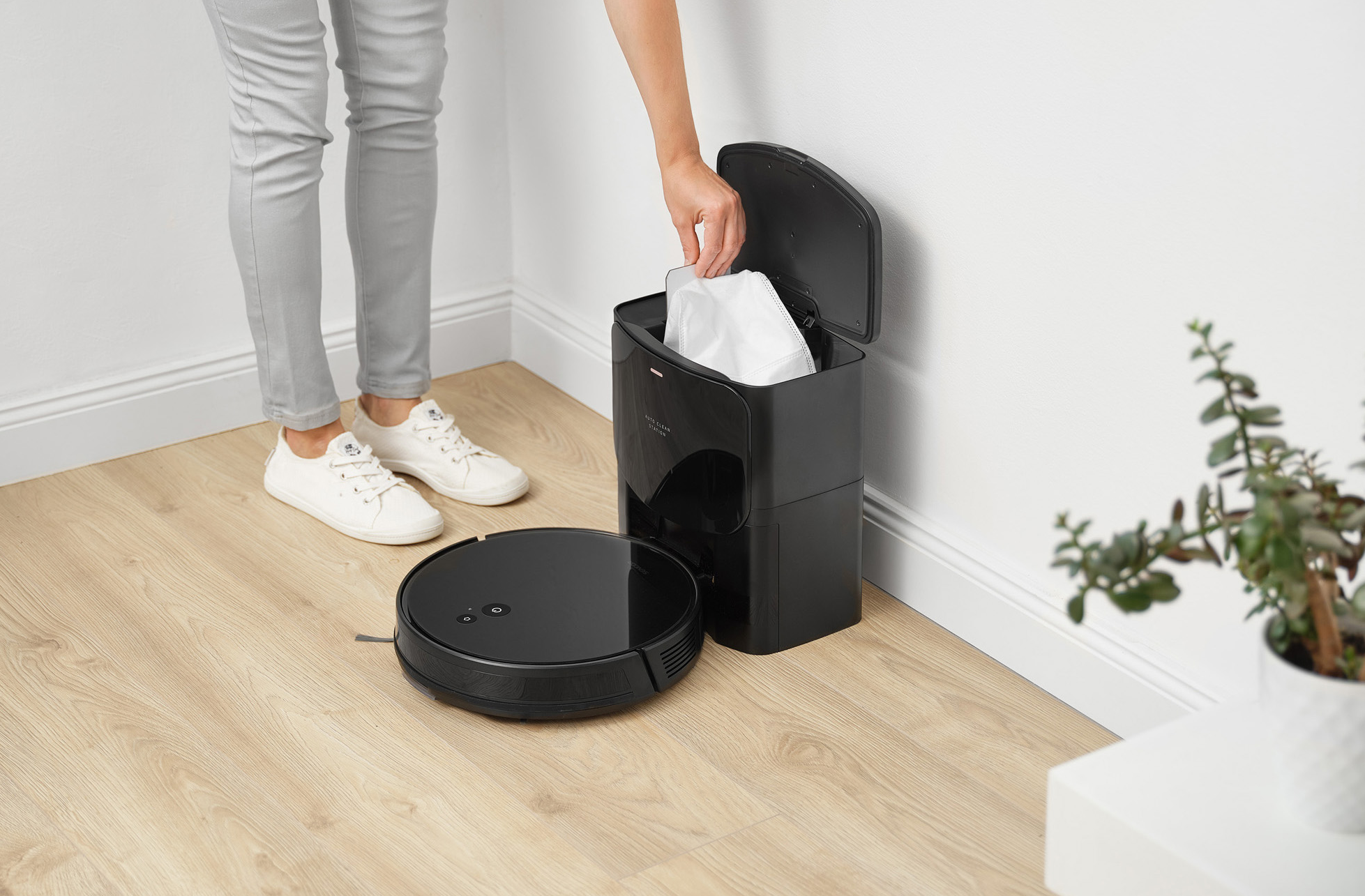 Egy személy egy zsákot helyez egy fekete porszívó portartójába. Egy fekete robotporszívó a padlón.