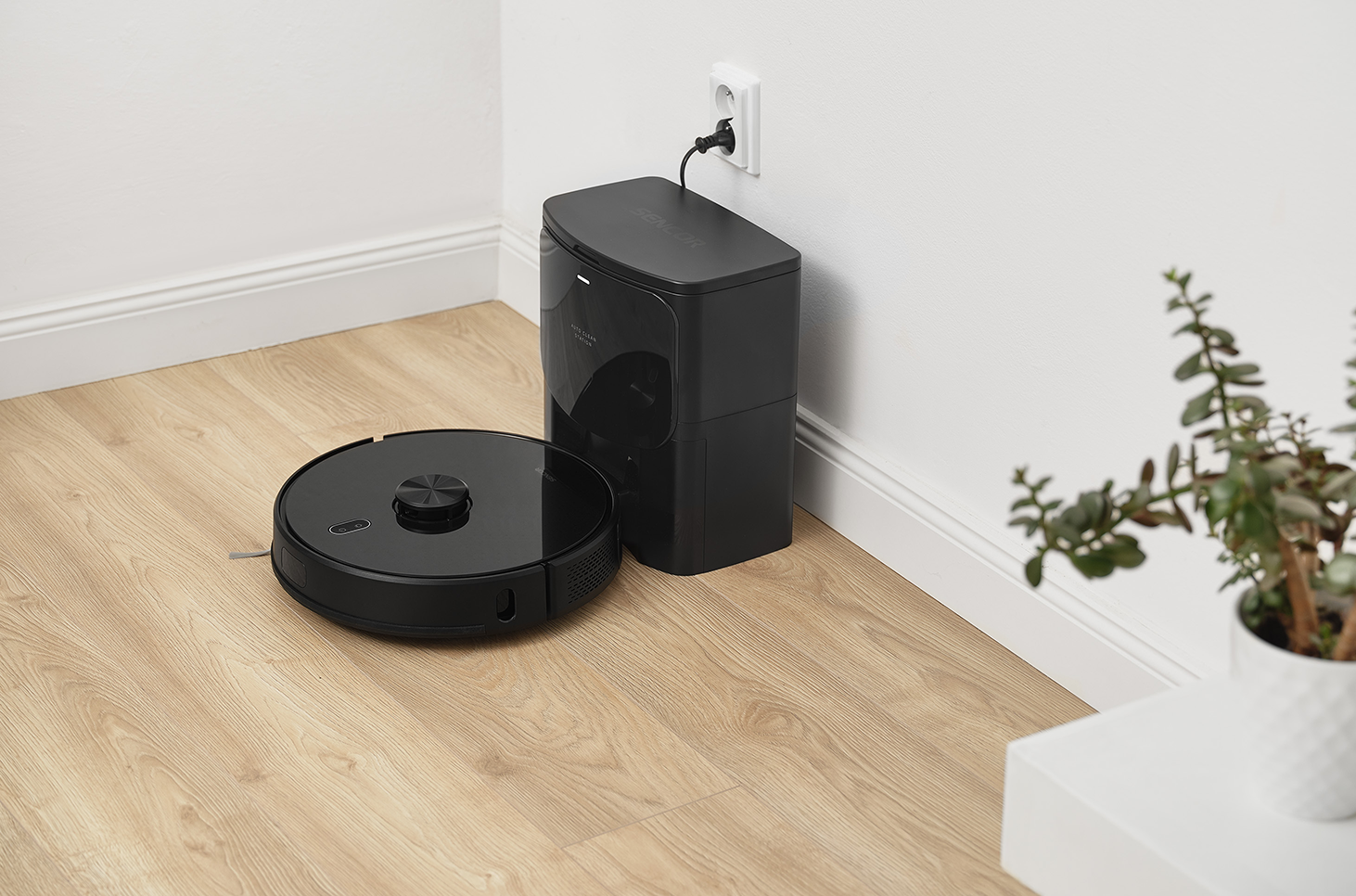 Robot porszívó töltőállomással, fehér fal és egy növény mellett.