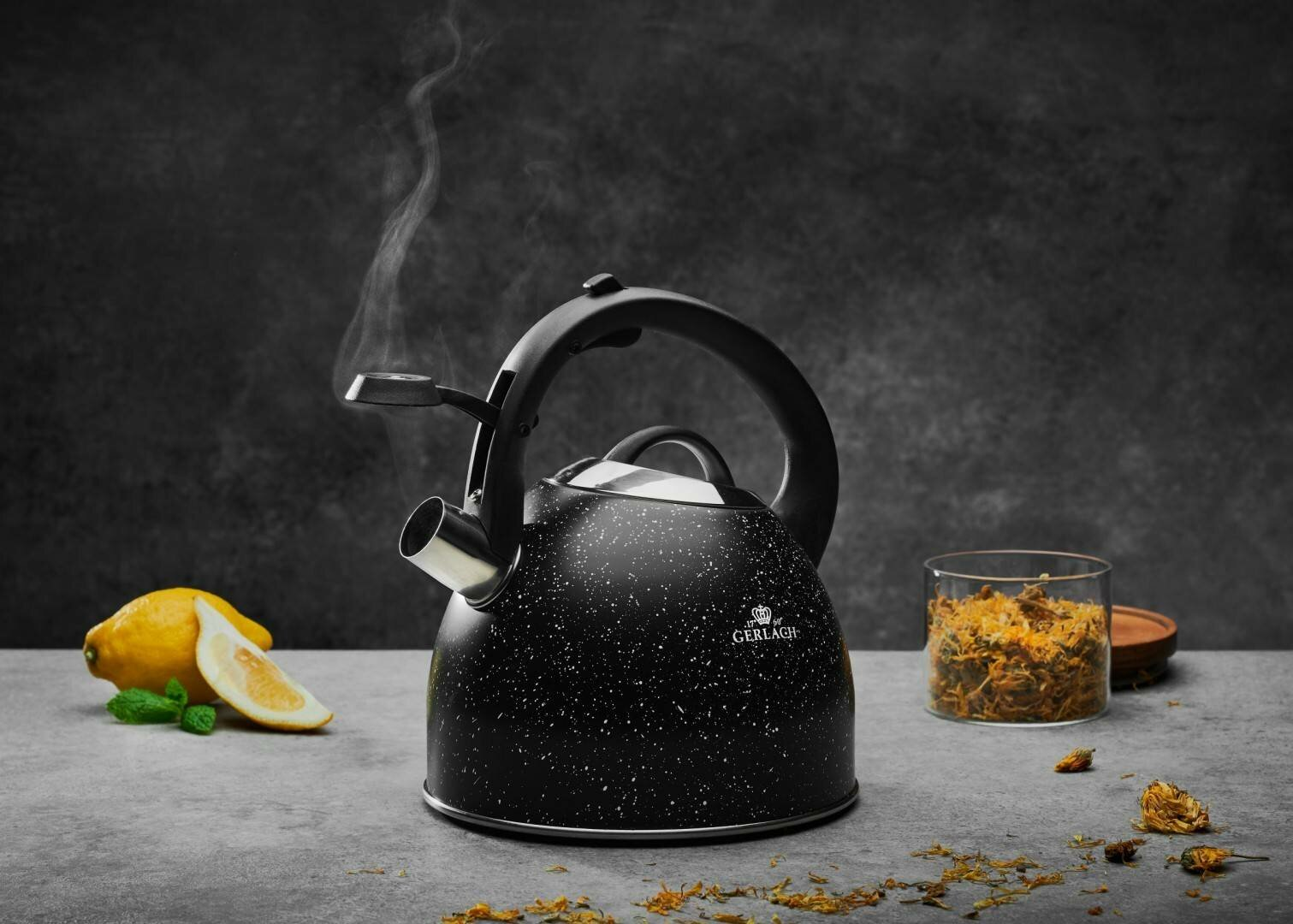 Czarny i srebrny czajnik z czarnym uchwytem. Para wydobywa się z dzióbka. Cytryna i zioła są w pobliżu.