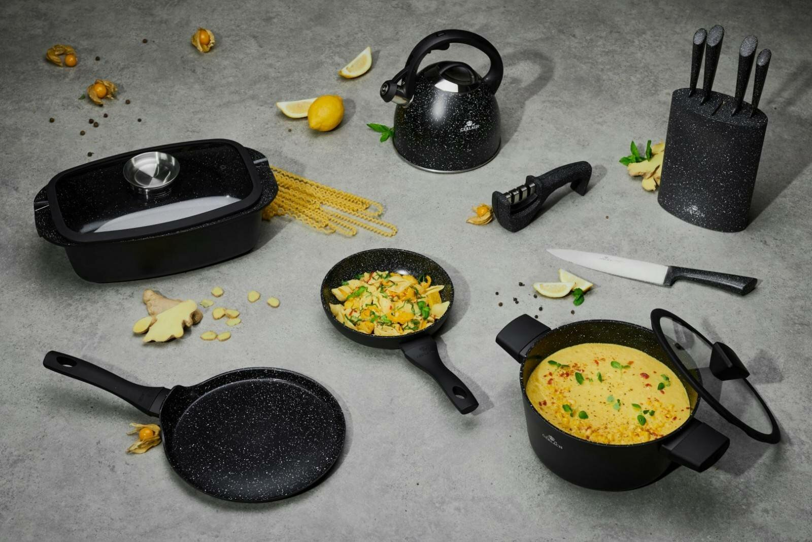 Narzędzia kuchenne: patelnie, garnek, blok na noże, czajnik i naczynie do pieczenia. Rozrzucone jedzenie i cytryny.