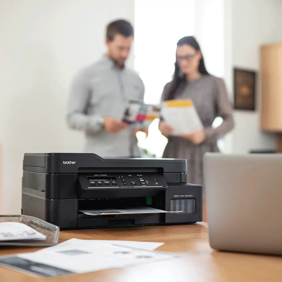 Scena biurowa z drukarką, laptopem i dwiema osobami.